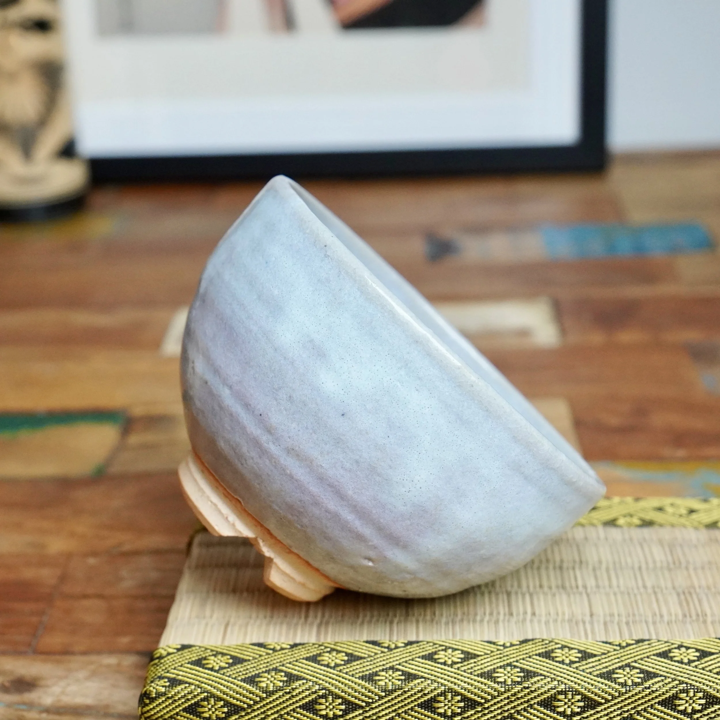 Moriwaki Fumitada's Hagi-yaki chawan, tilted to show its pale blue-grey glaze, lilac tones, and unglazed light orange foot on a woven mat.