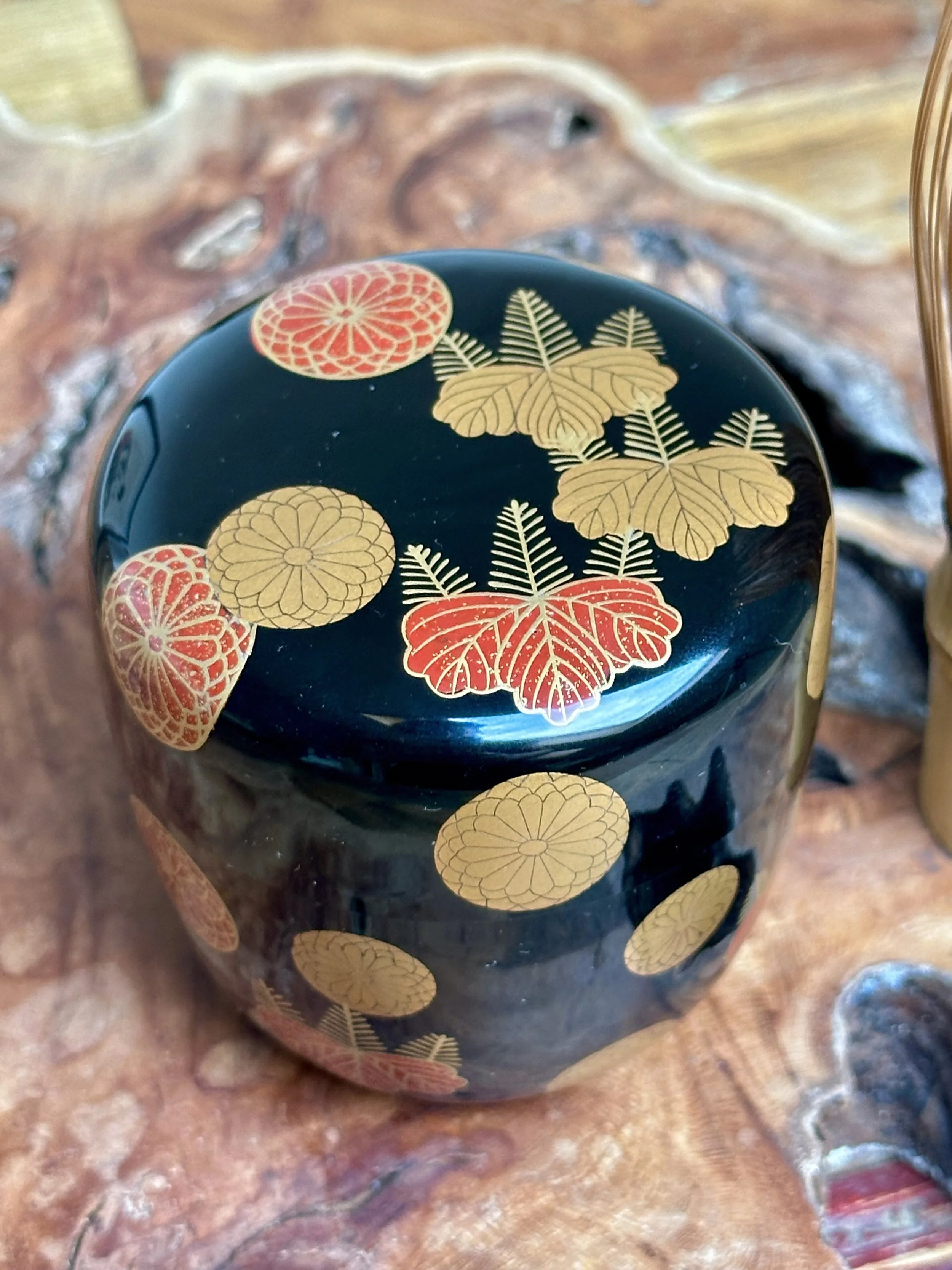 Black lacquered natsume tea caddy by Yūsai, featuring scattered gold chrysanthemum and red paulownia crests, reflecting light.