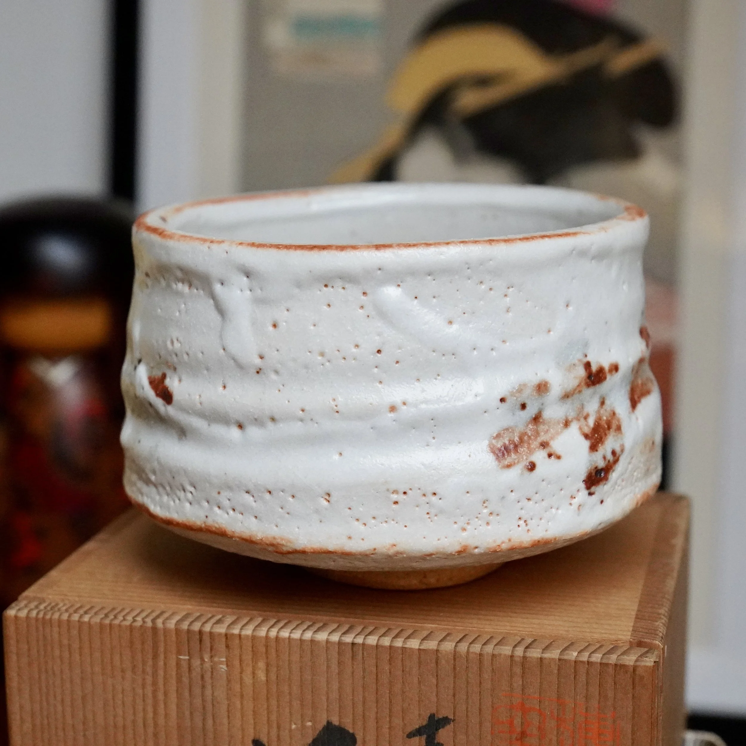White chawan with pitted texture and rust-orange glaze accents, resting on a finger-jointed wooden tomobako with dark script.