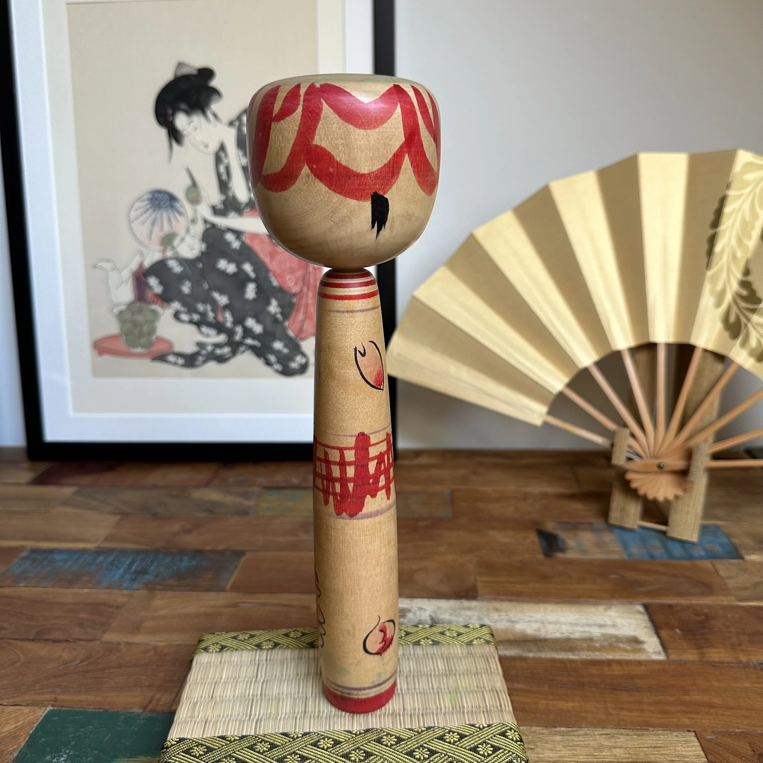 Seya Jūji's Nakanosawa Takobozu kokeshi, a wooden doll with a large red-patterned head and black mark. Its body has red stripes and stylized blossoms, on a woven mat.