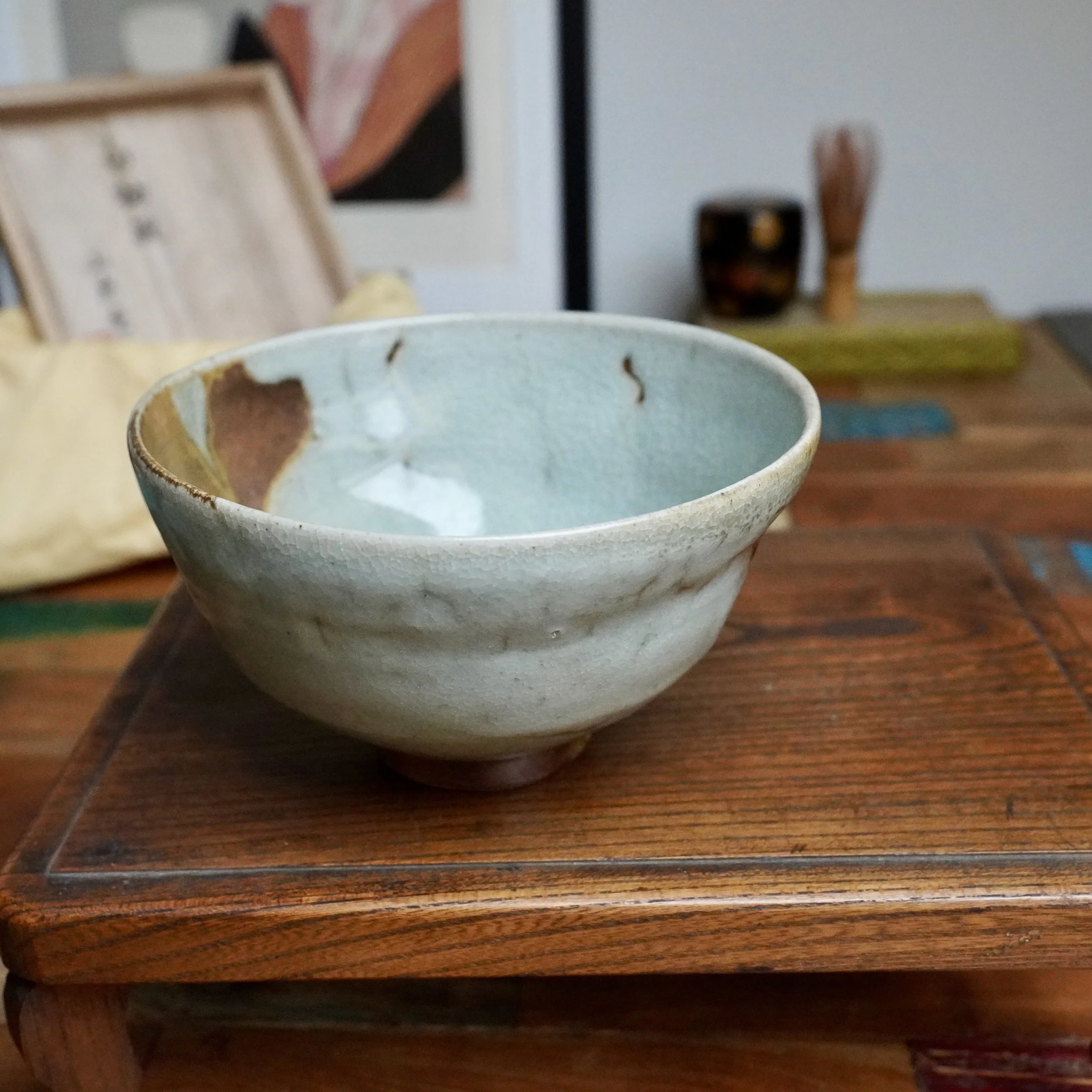 Sansōan chawan, pale blue-grey ice crackle glaze flowing to rust-brown and ochre, on a wooden surface with its tomobako behind.