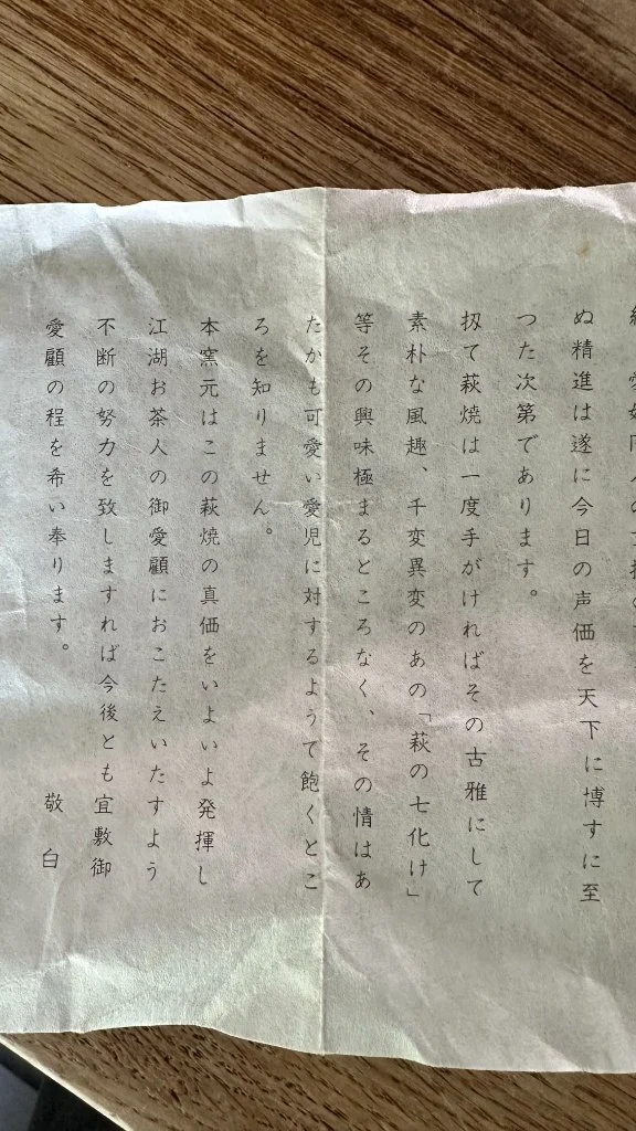 Vertically folded, crumpled off-white shiori document signed by Hirose Tanga, featuring black Japanese text, resting on a wooden surface.