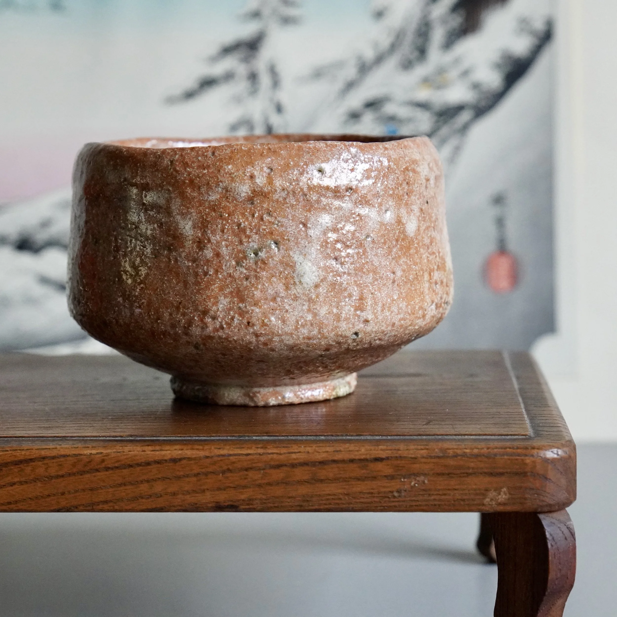 Aka-raku chawan by Sawada Hiroyuki, a rugged reddish-brown bowl with pitted texture and soft white glaze drips, resting on a wooden stand.