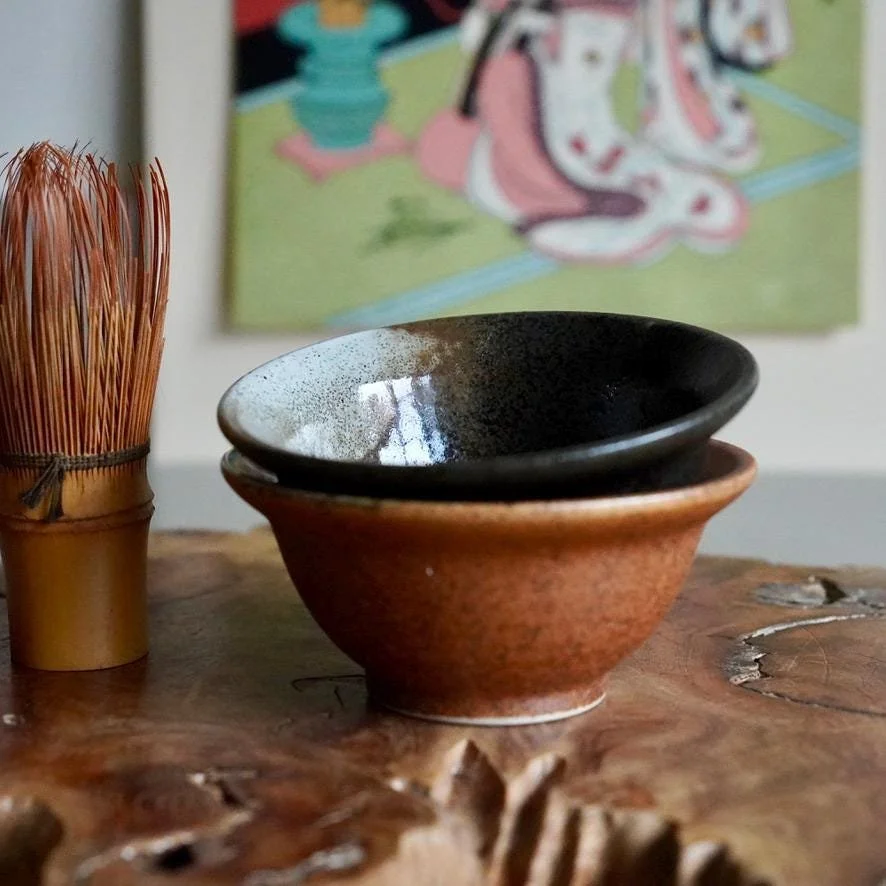 Temmoku ware iron-glaze ceramic cup set deep brown glaze Kyoto, Shōwa period Japan, displayed stacked on wooden plank next to chasen with ukiyoe print on background