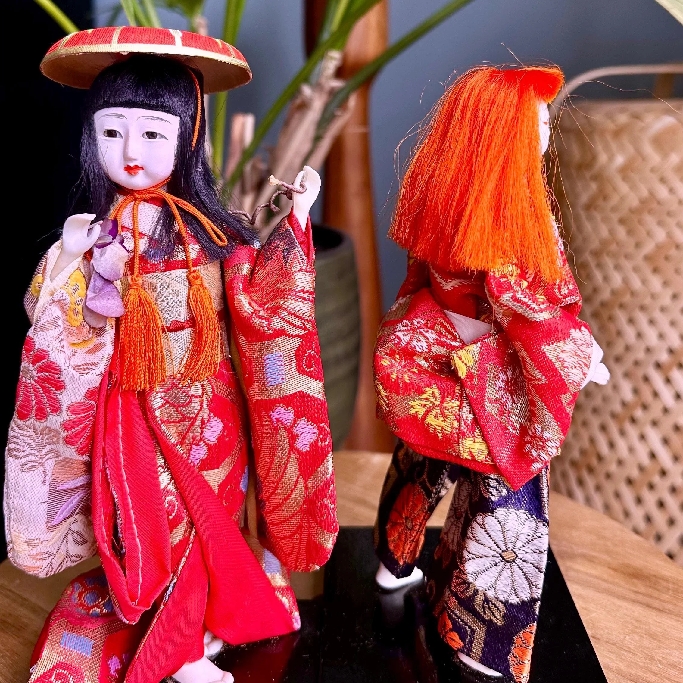 Kabuki theater ningyo dolls in red and gold kimonos on black bases. Left doll with black hair and red hat faces forward; right doll with wild orange hair is turned away.
