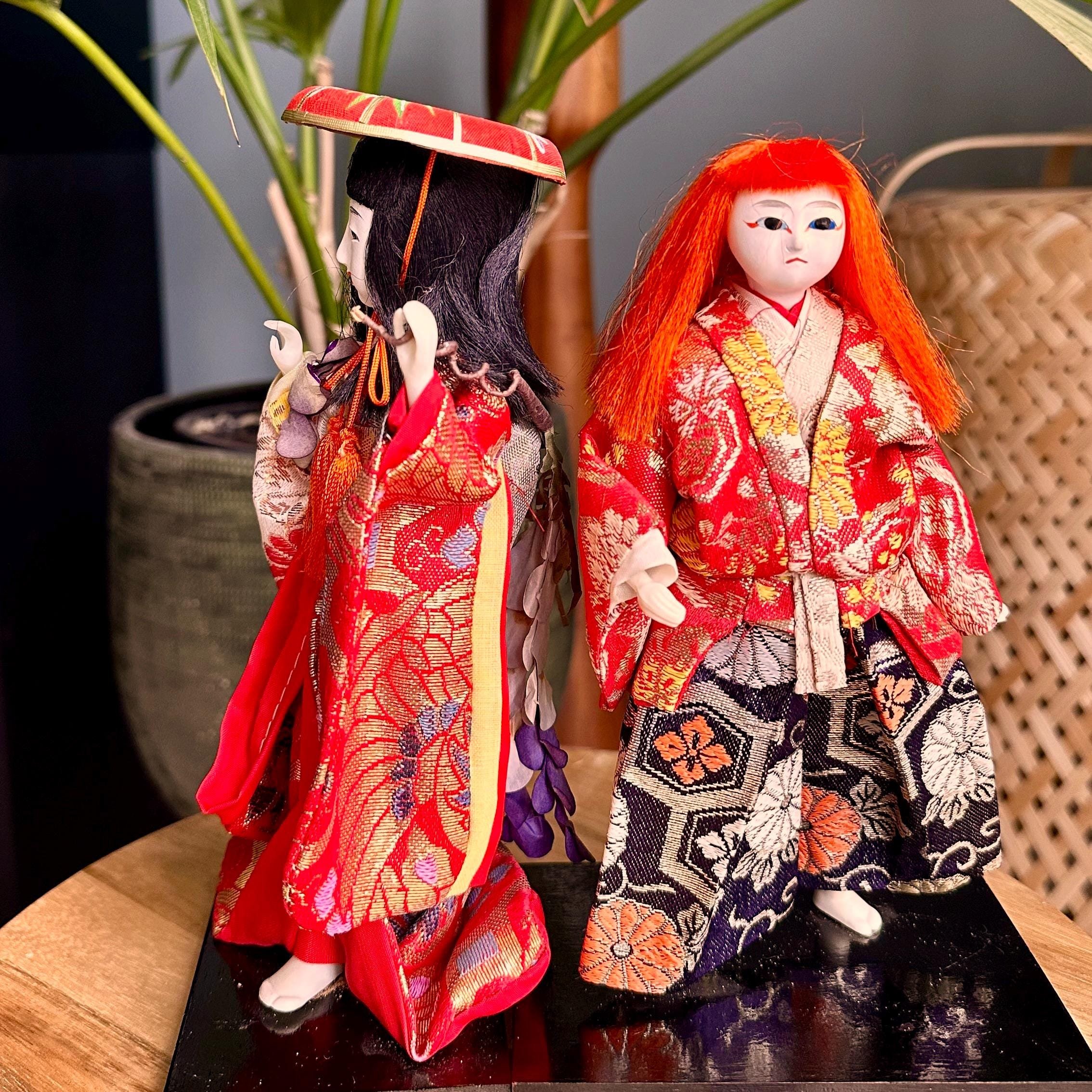 Pair of Kabuki theater ningyo dolls in red and gold patterned kimonos on black bases. The left doll has black hair and a red hat, the right has wild orange-red hair.