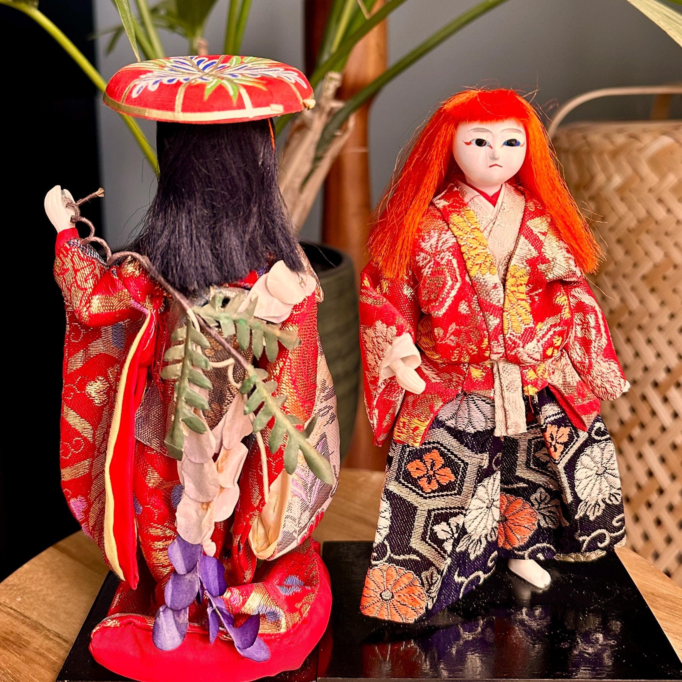 Two Kabuki Kagamijishi dolls: a composed lady Yayoi with black hair and red hat, and a dynamic lion spirit with wild orange hair. Both wear red and gold kimonos on black bases.