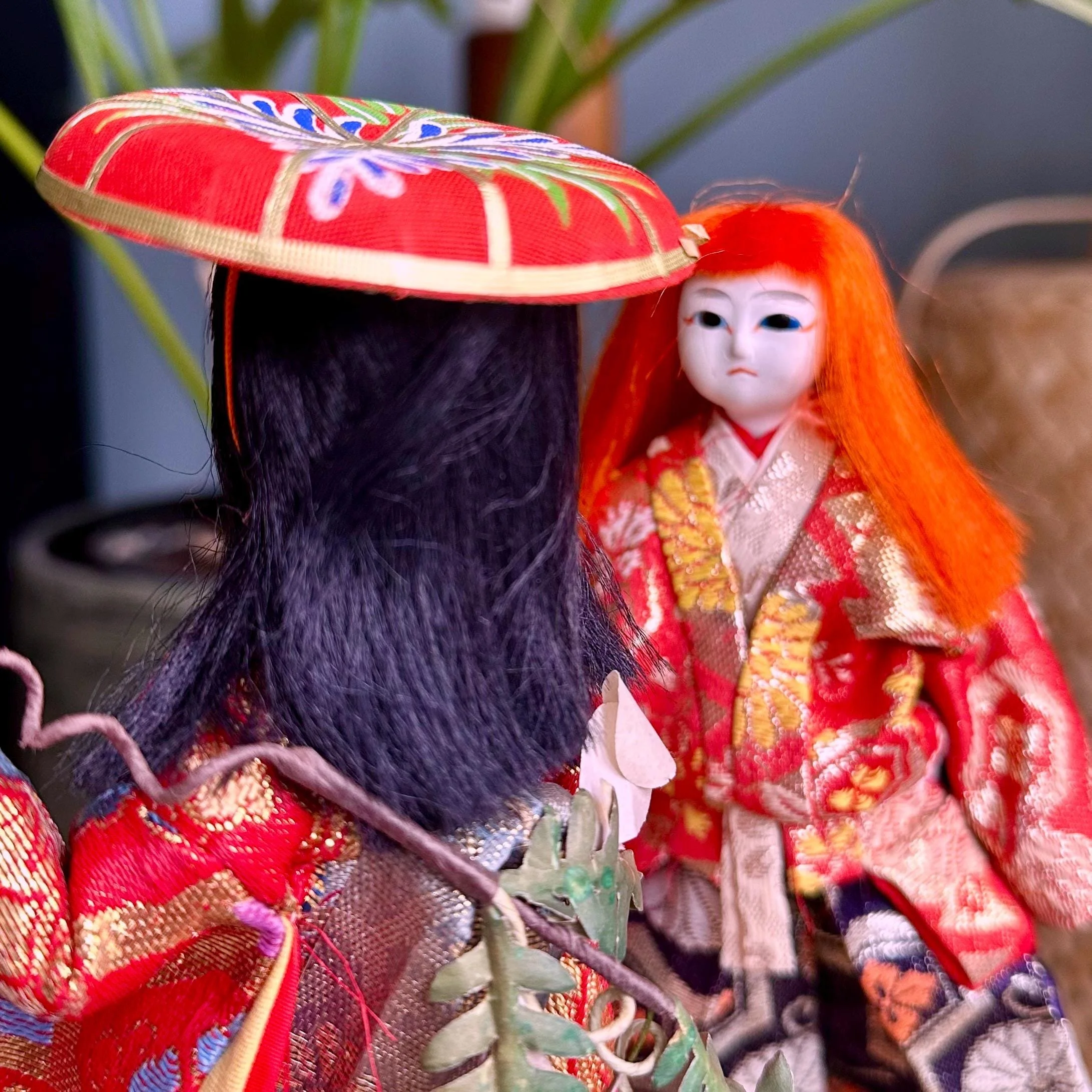 Close-up of two Kabuki ningyo dolls. Left, black hair, red ceremonial hat with gold trim, red & gold robe with foliage detail. Right, white face, blue eyes, orange hair, red and gold patterned kimono.