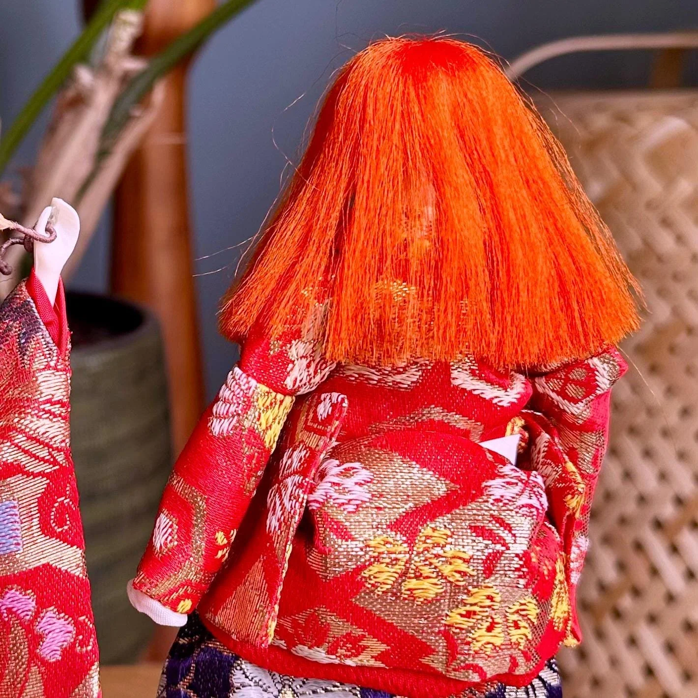 Rear and side view of a Kabuki doll with wild orange-red hair, wearing a red kimono patterned with gold and white brocade. Second doll partially visible on left.