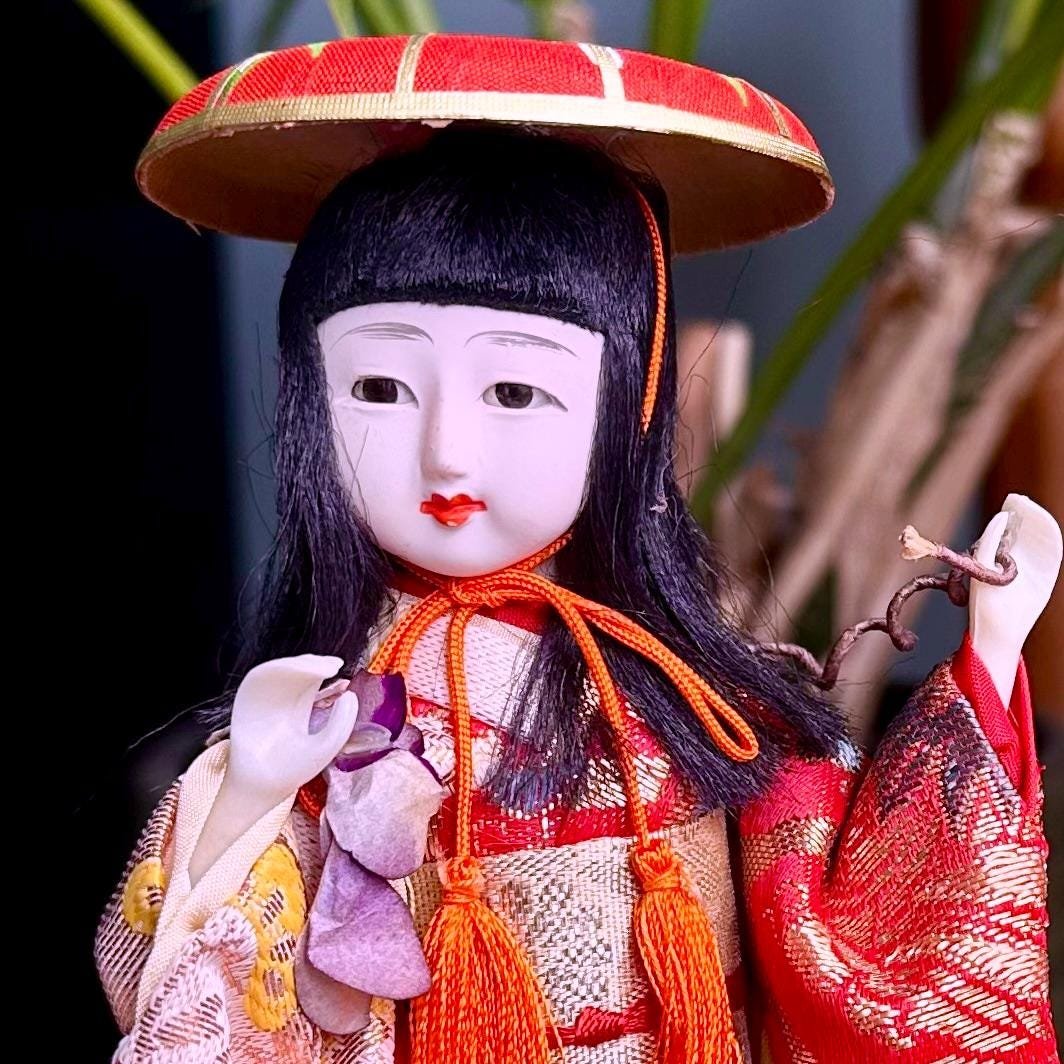 Front view of a Kabuki theater ningyo doll with a white gofun face, black hair, red ceremonial hat, and elaborate red and gold kimono, holding purple petals and a gnarled branch.