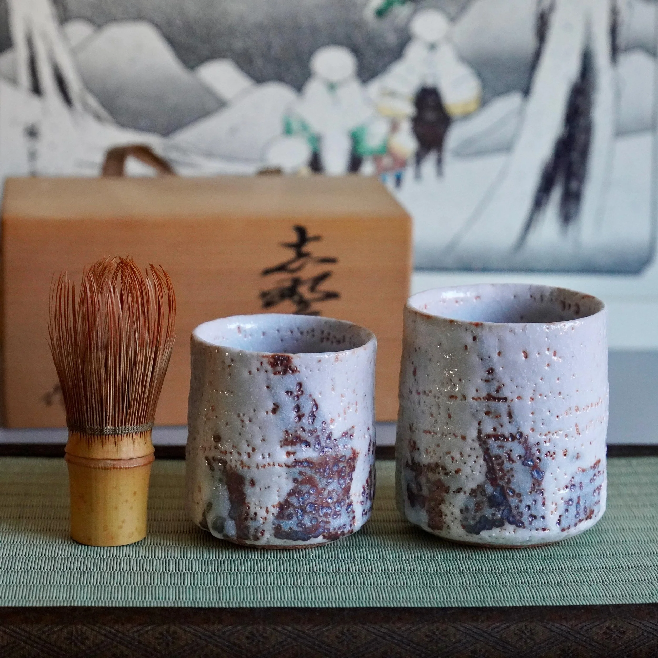 Two Shino yunomi cups with rough white feldspar glaze and reddish-brown spots, next to a bamboo whisk and a wooden box signed by Yōkun, on a tatami mat.