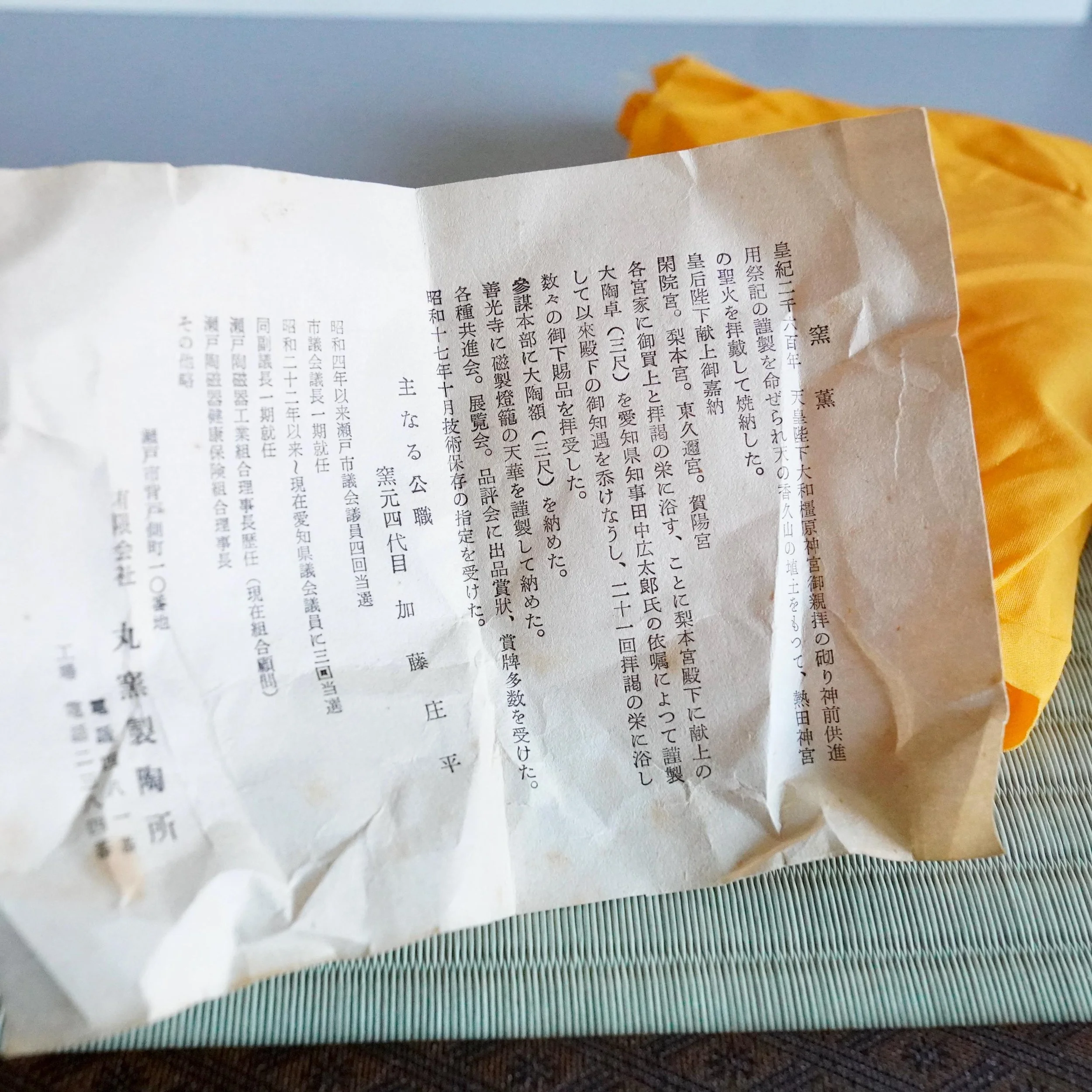 Marugama kiln's printed history, a folded document with dense Japanese text, resting on a green striped mat with yellow fabric behind.