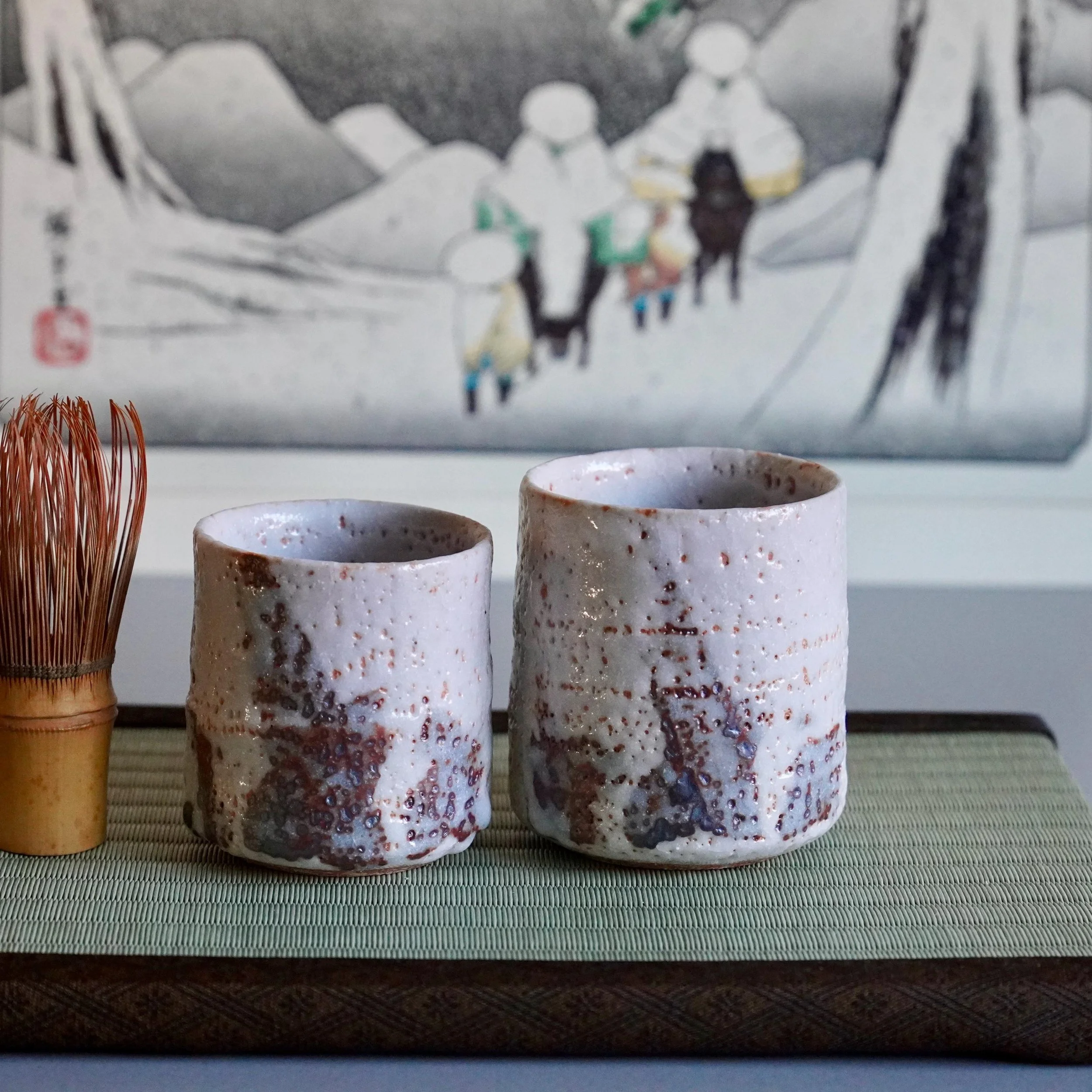 Katō Shōhei's two Shino yunomi, one larger, featuring textured white and reddish-brown glaze, beside a bamboo matcha whisk.