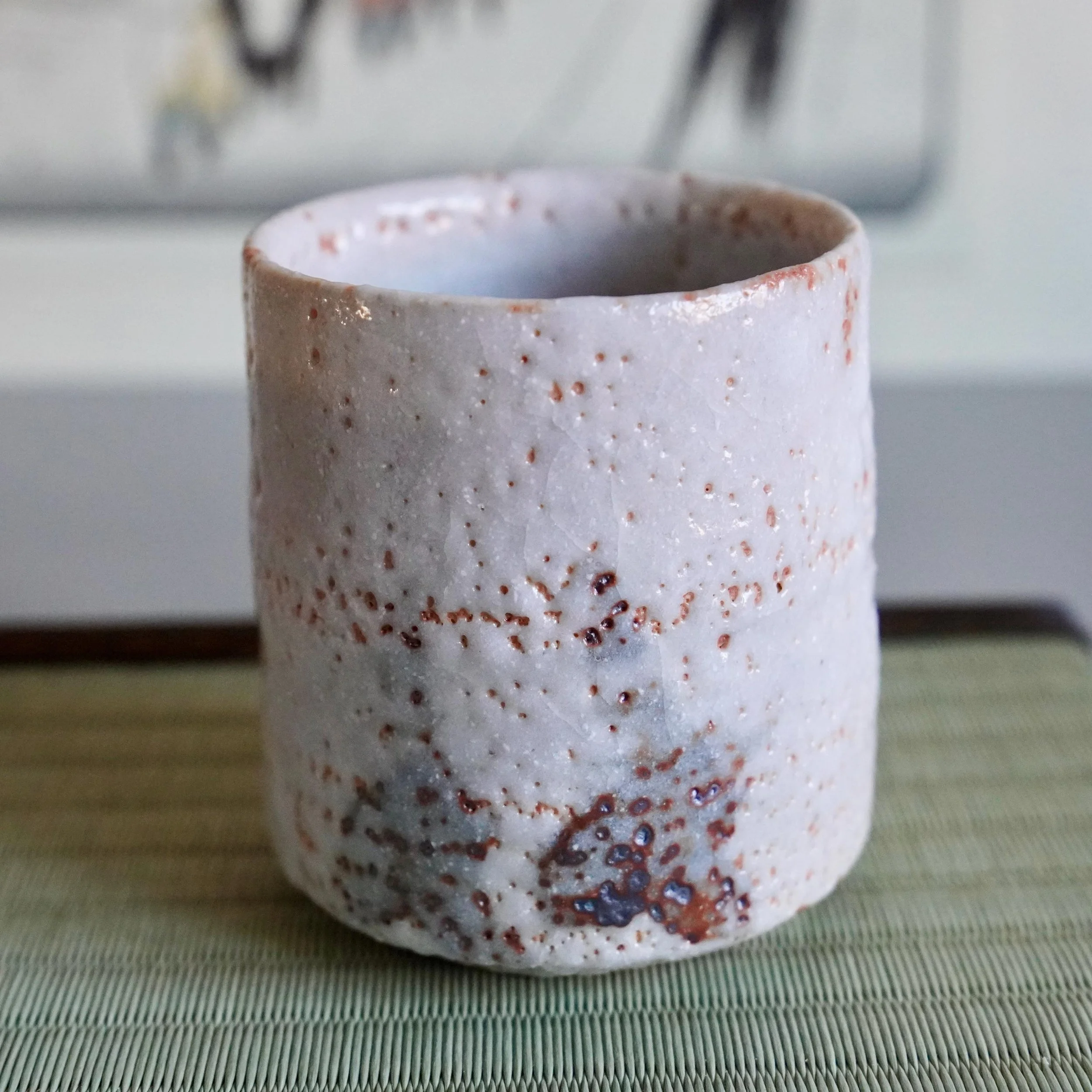 Shino yunomi by Katō Shōhei (Yōkun) with white feldspar glaze, rough patches, and reddish-brown and dark iron spots, on tatami mat.