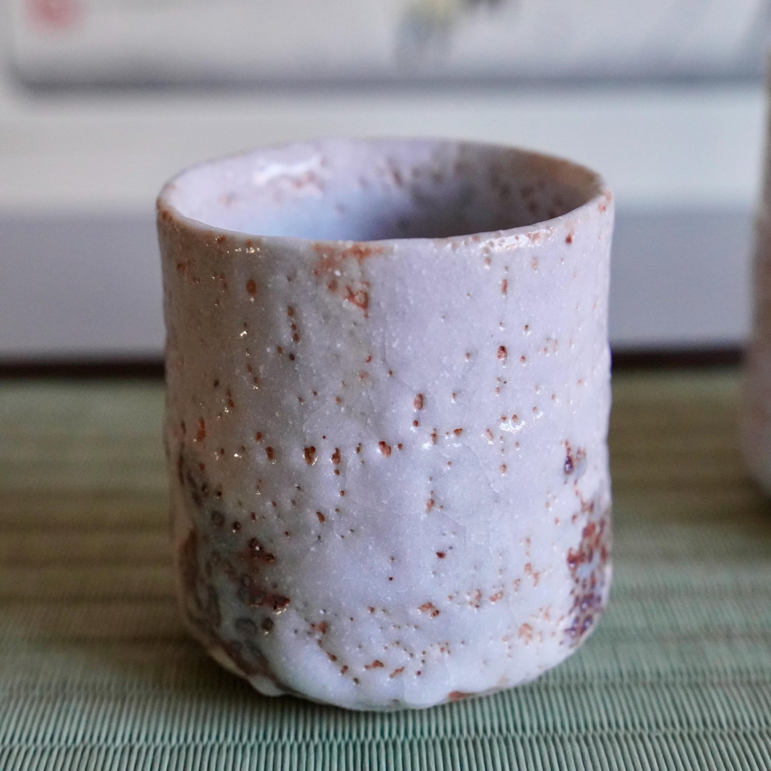 Shino yunomi with a thick, textured purplish-white feldspar glaze, revealing reddish-brown clay and dark iron spots, on a green ribbed mat, by Katō Shōhei (Yōkun).