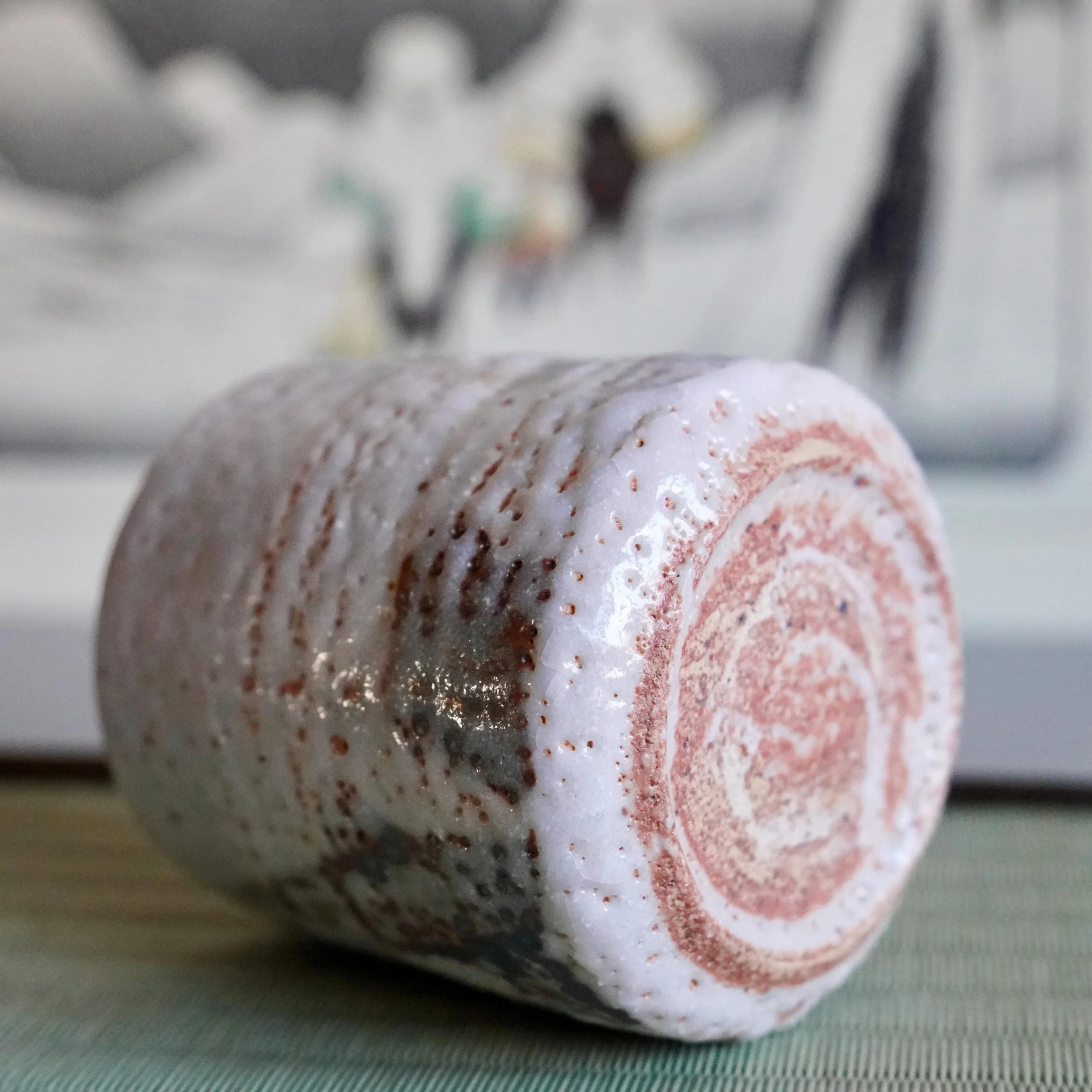 Yōkun Shino yunomi, on its side, with thick white textured glaze, reddish-brown showing through, dark iron spots, and a spiral pattern on the base.
