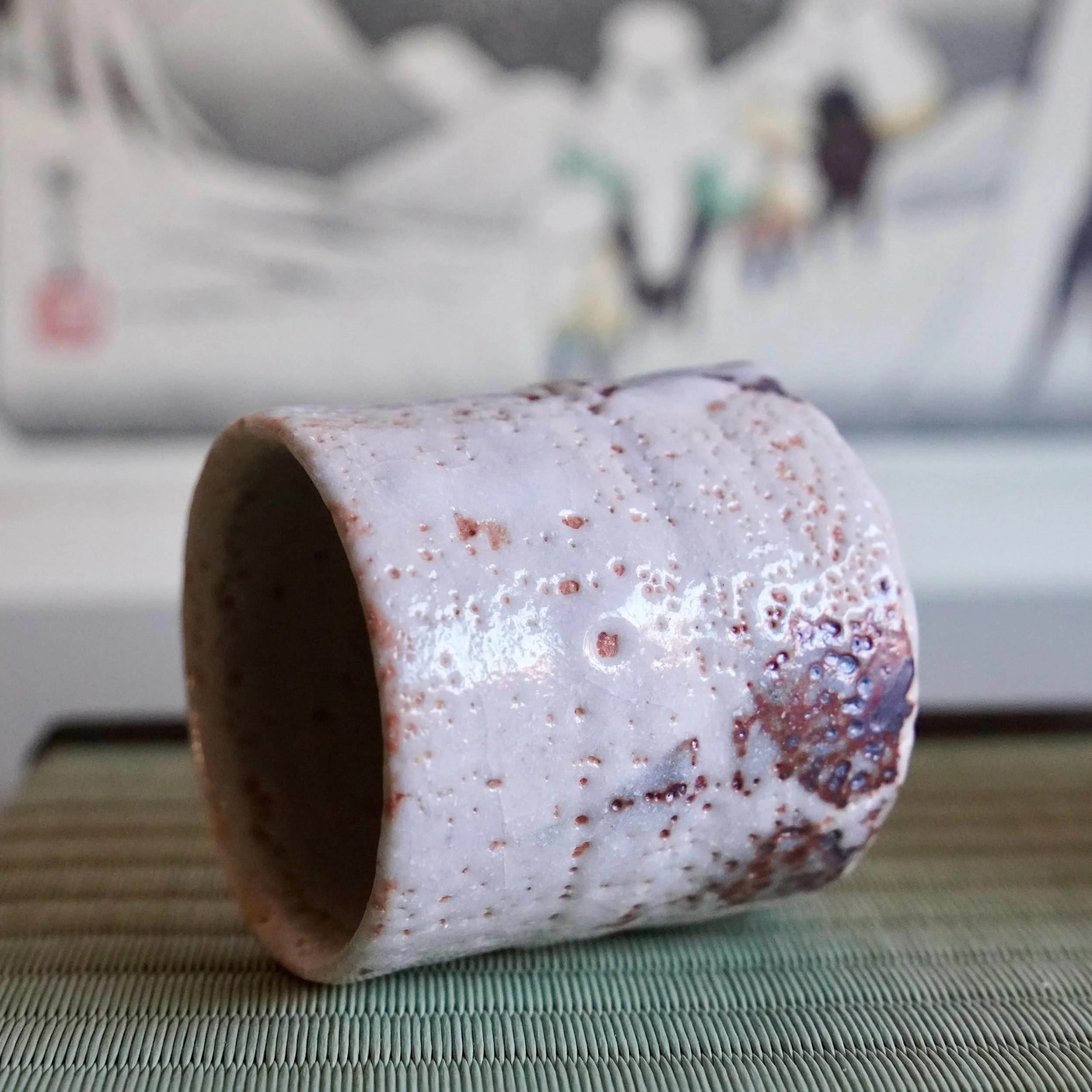 Shino yunomi lying horizontally, with a textured white feldspar glaze, reddish-brown patches, dark iron spots, and a pitted, volcanic surface on a woven mat.