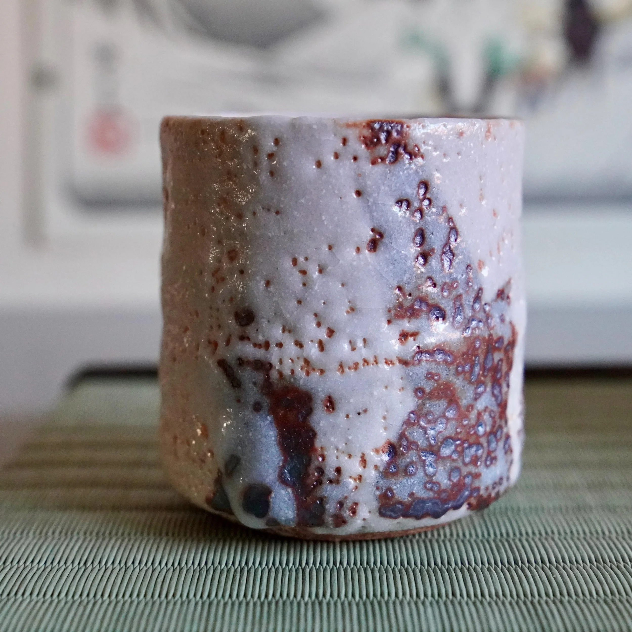 Katō Shōhei (Yōkun) Shino yunomi with thick white feldspar glaze, reddish-brown sections, and dark iron spots, on a green tatami mat.