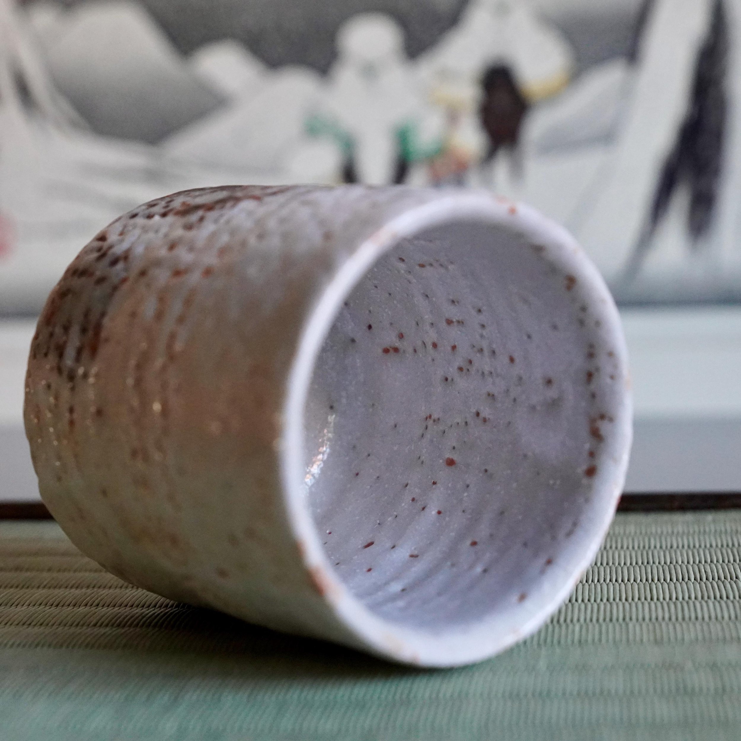 Shino yunomi cup with speckled white and reddish-brown glaze, lying on its side on a green tatami mat.