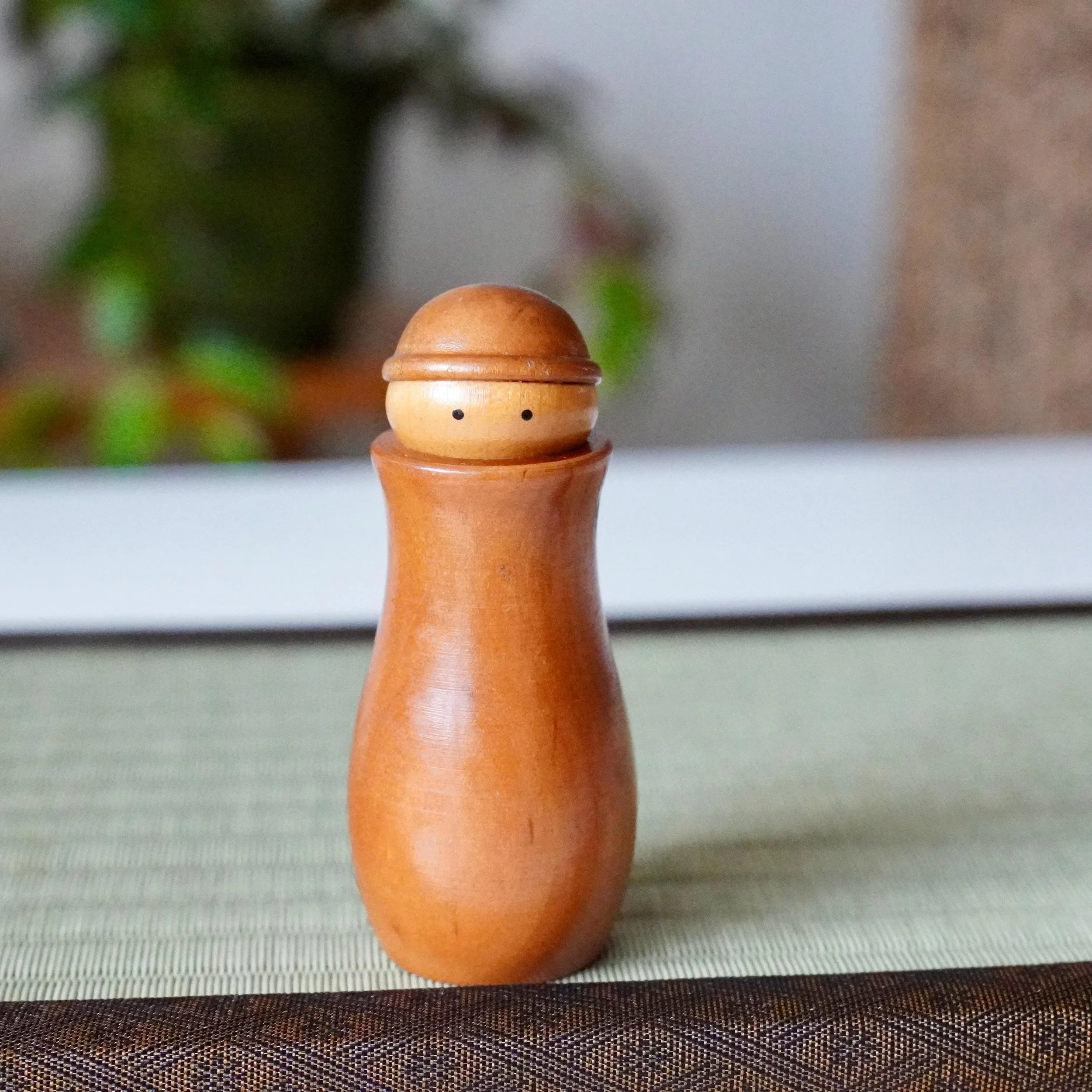 Kōkyū wooden Kokeshi toothpick holder, front view. It's light brown with a smooth, polished finish and two black eyes, sitting on a woven mat with a dark border.