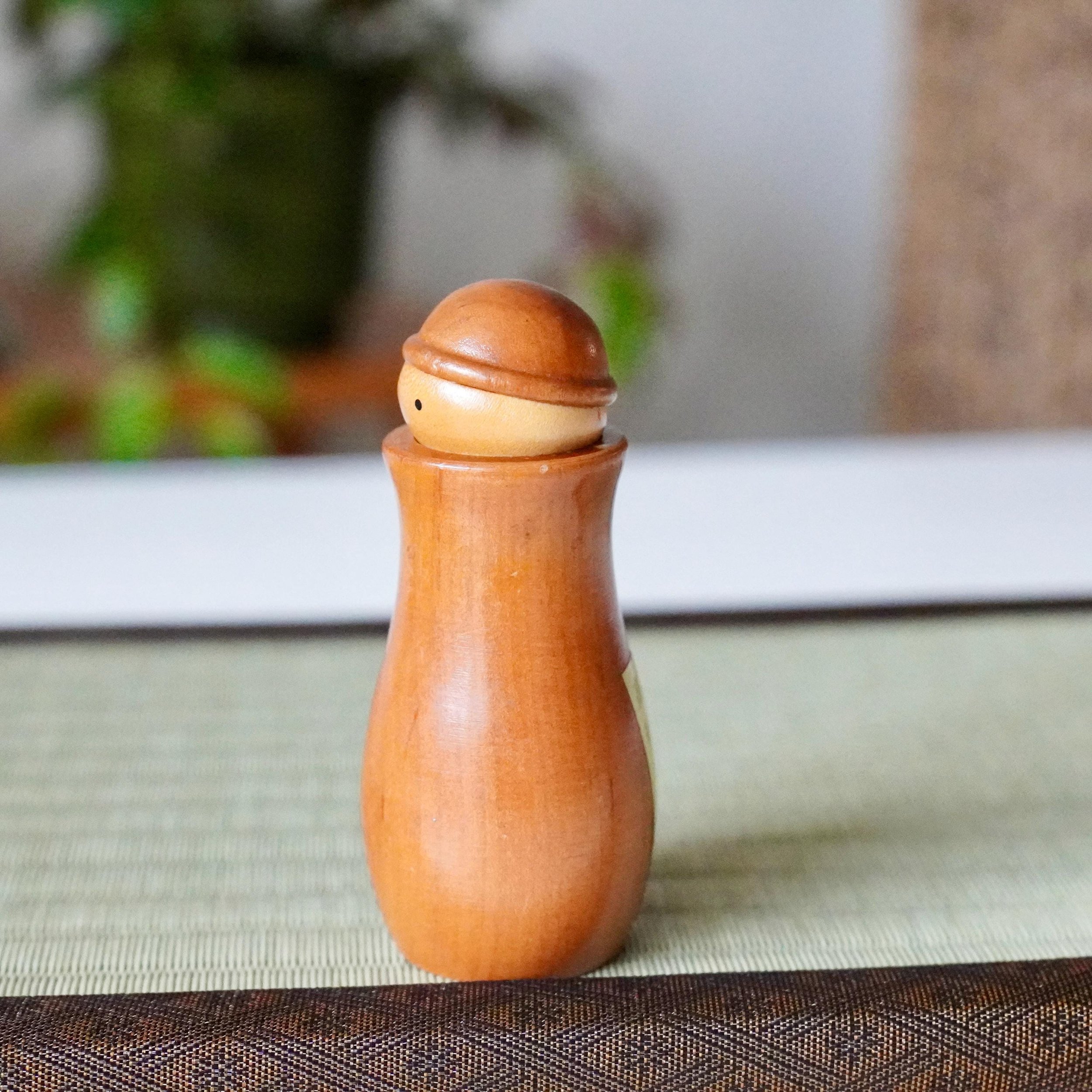 Kōkyū kokeshi toothpick holder, smooth light brown wood, with a single black eye, displayed on a textured tatami mat.