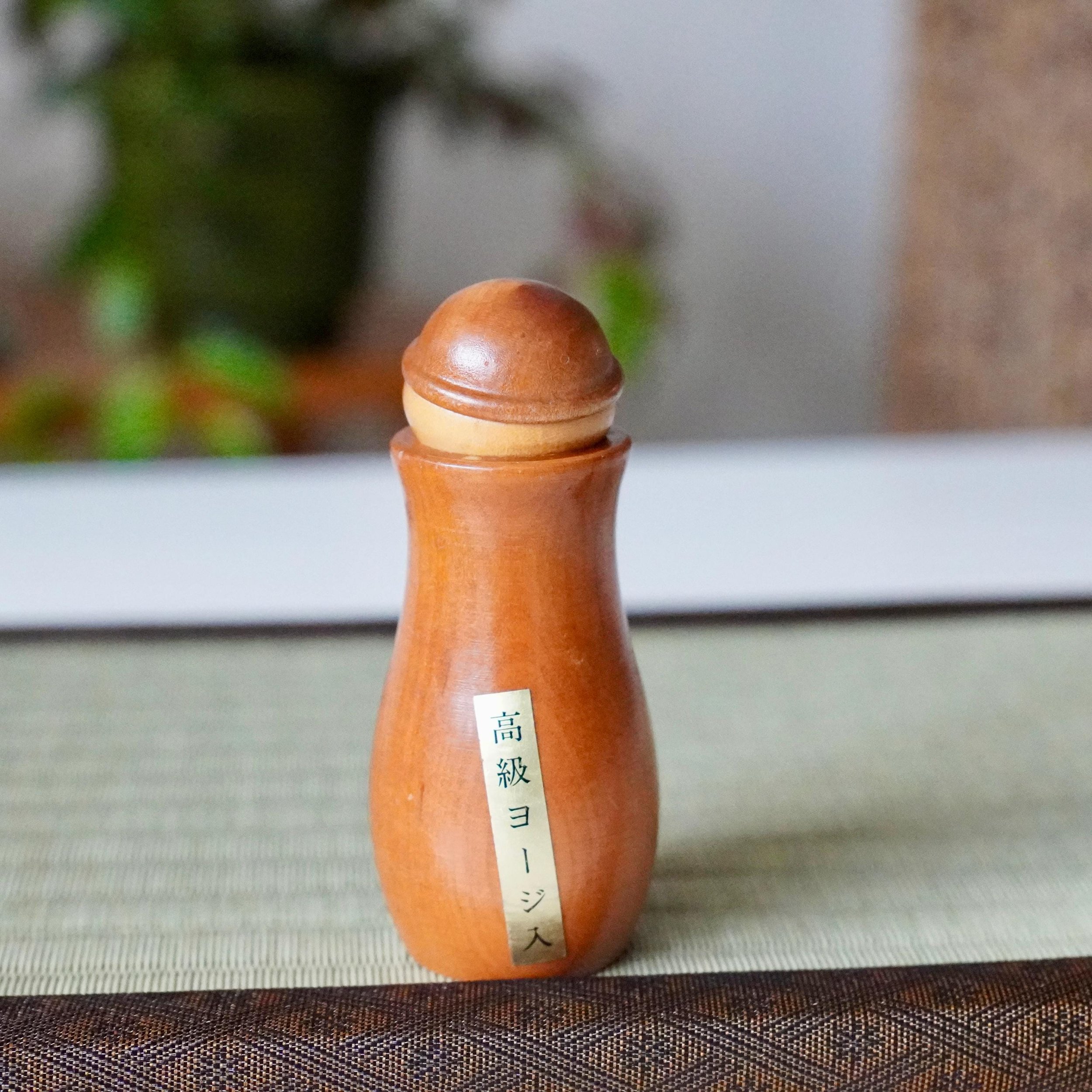 Smooth, reddish-brown wooden kokeshi toothpick holder, front view, with a Kōkyū label, resting on a woven mat. From the Okazaki Museum.