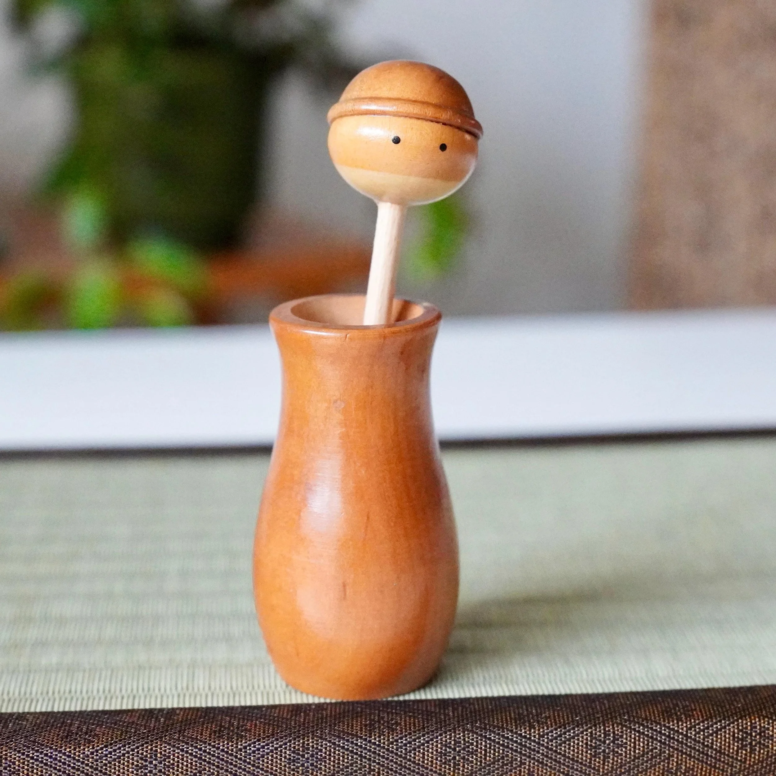 Kōkyū wooden kokeshi toothpick holder with a spherical head featuring two black dots for eyes and a carved ridge, sitting in its base on a tatami mat.