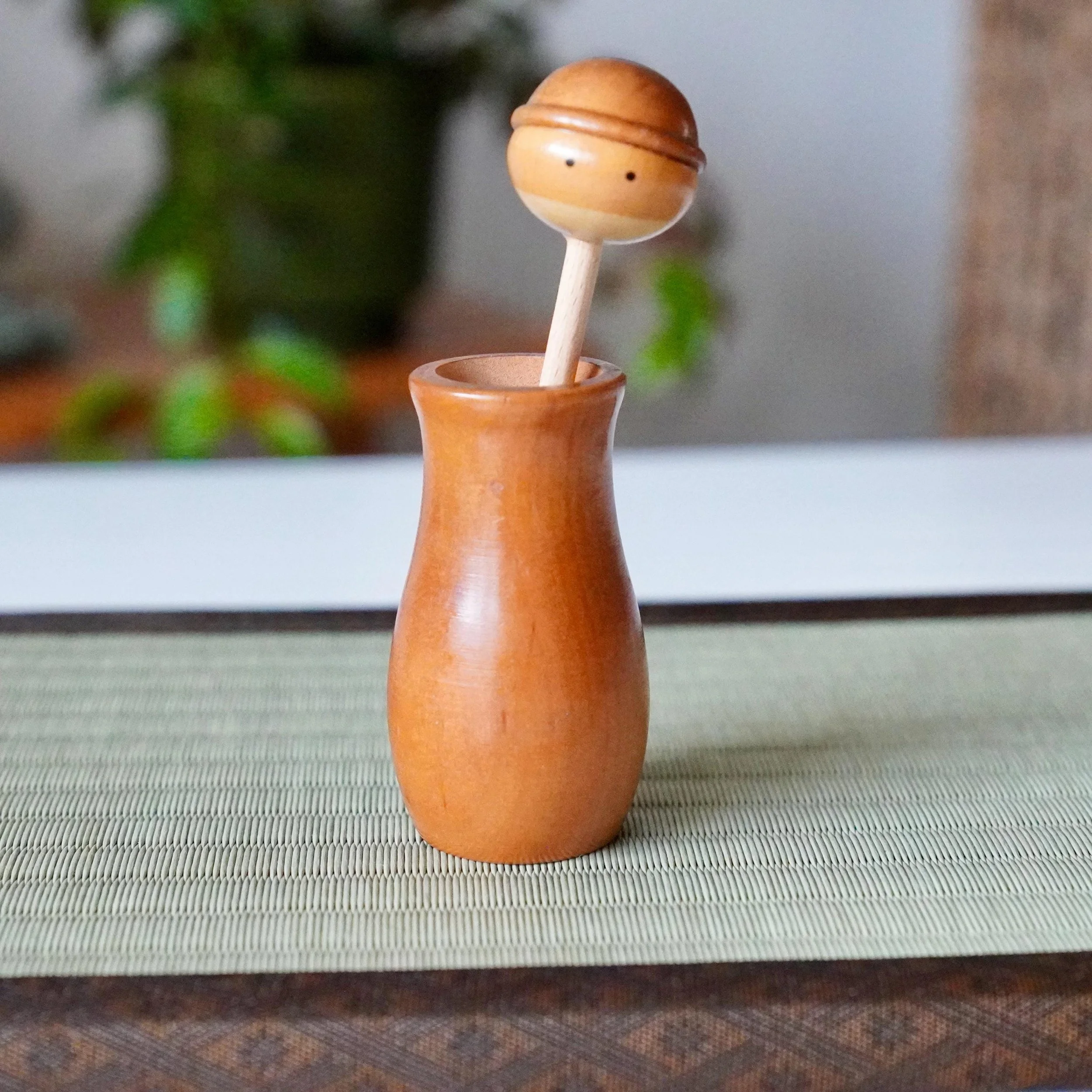 Kōkyū wooden kokeshi toothpick holder: a round head with simple eyes and band detail on a stick, inserted into its polished reddish-brown vase-shaped body.