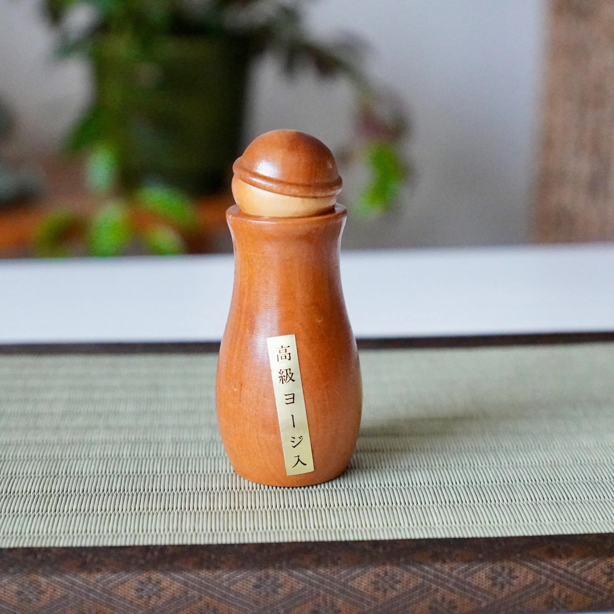Smooth wooden kokeshi toothpick holder with a rounded head, a gold label inscribed 'Premium Toothpick Container,' on a tatami mat.