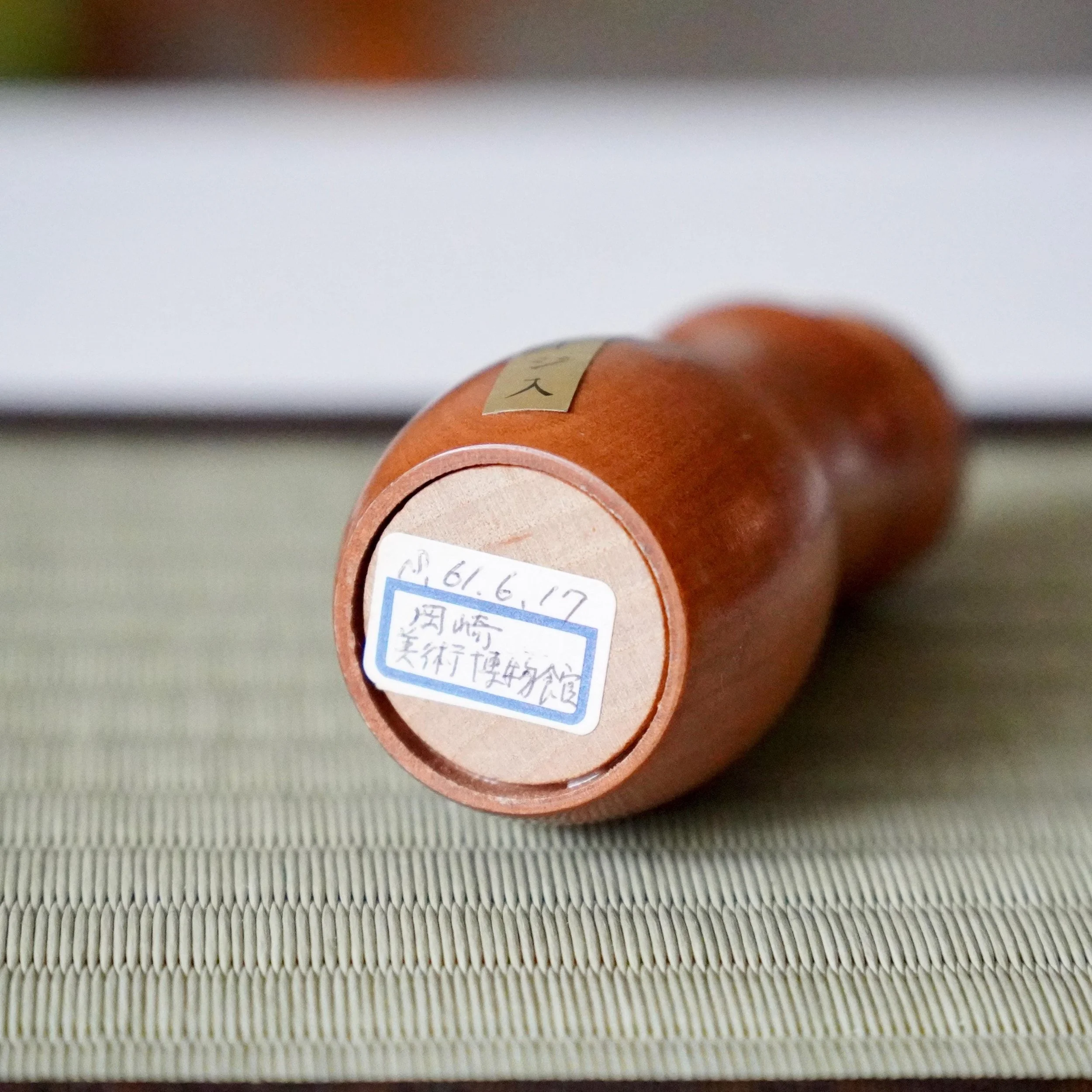 Underside view of a light brown wooden kokeshi with a white sticker dated S. 61.6.17 and "岡崎 美術博物館" (Okazaki Museum of Art) in blue ink.