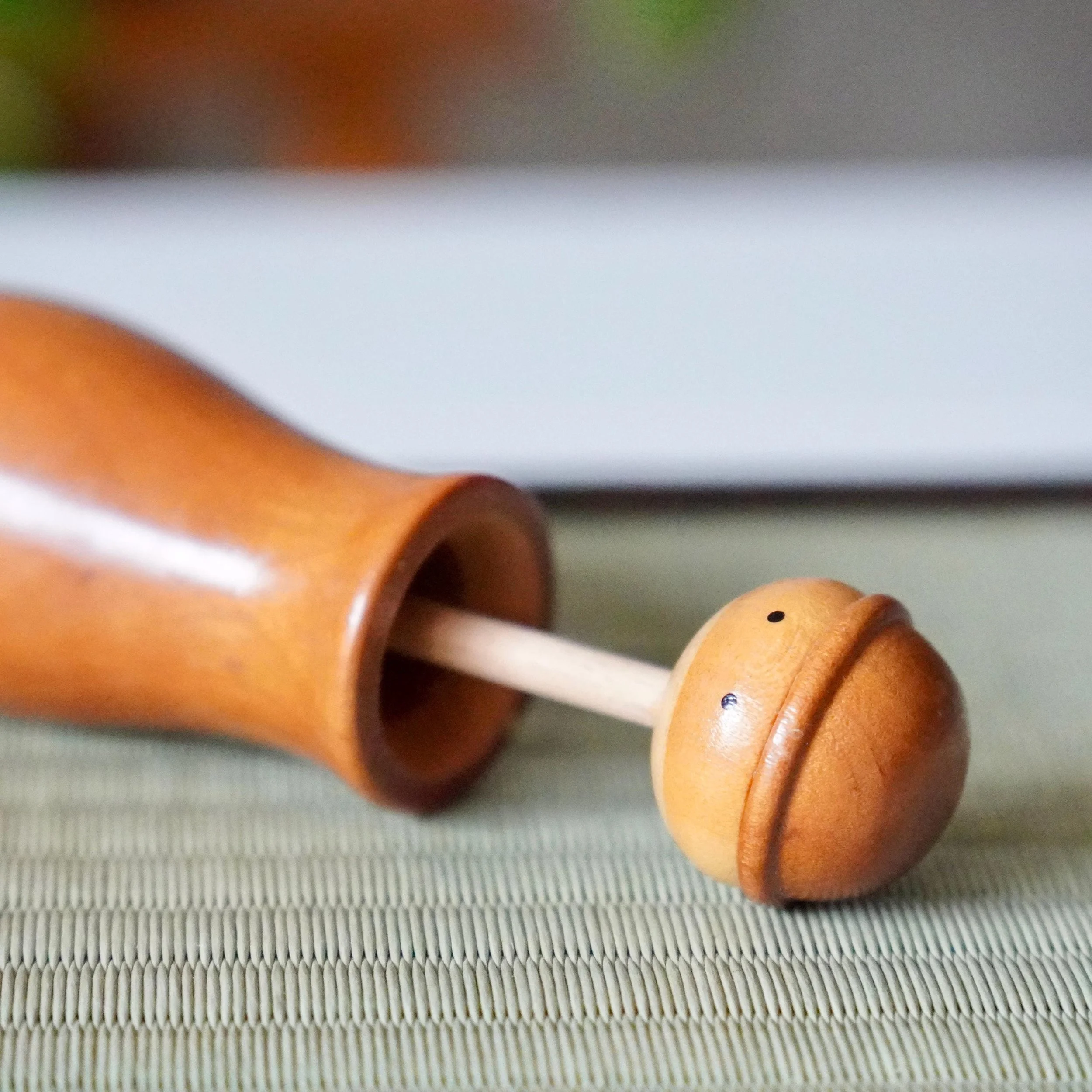 Kōkyū kokeshi toothpick holder, wooden, lying on a ribbed mat with its rounded head lifted to reveal a pale stem. Small black dots mark the head.