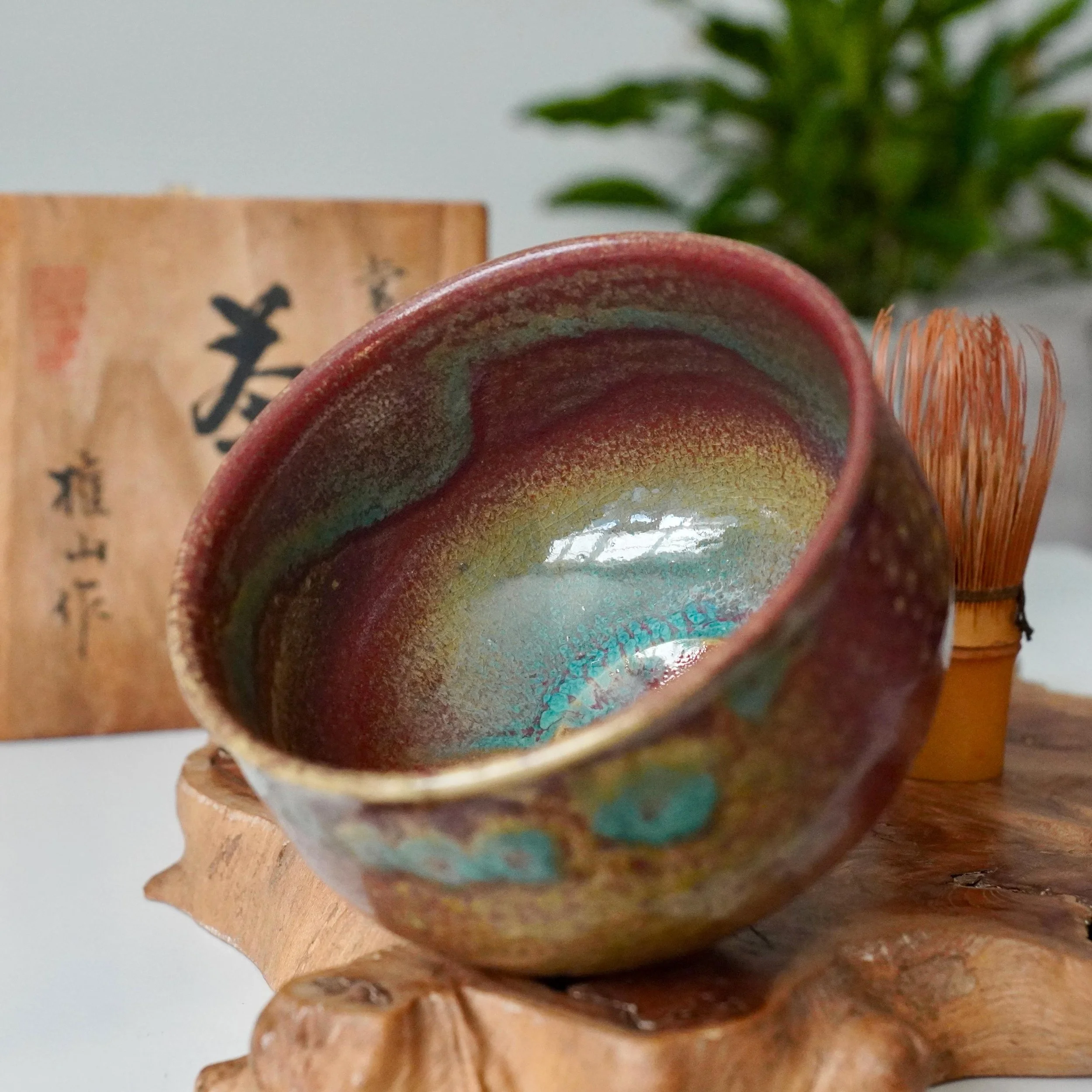 Kenzan chawan featuring dynamic yōhen glaze in ruby red, gold, and turquoise, perched on a rustic wood stand. A chasen and tomobako box are in the background.