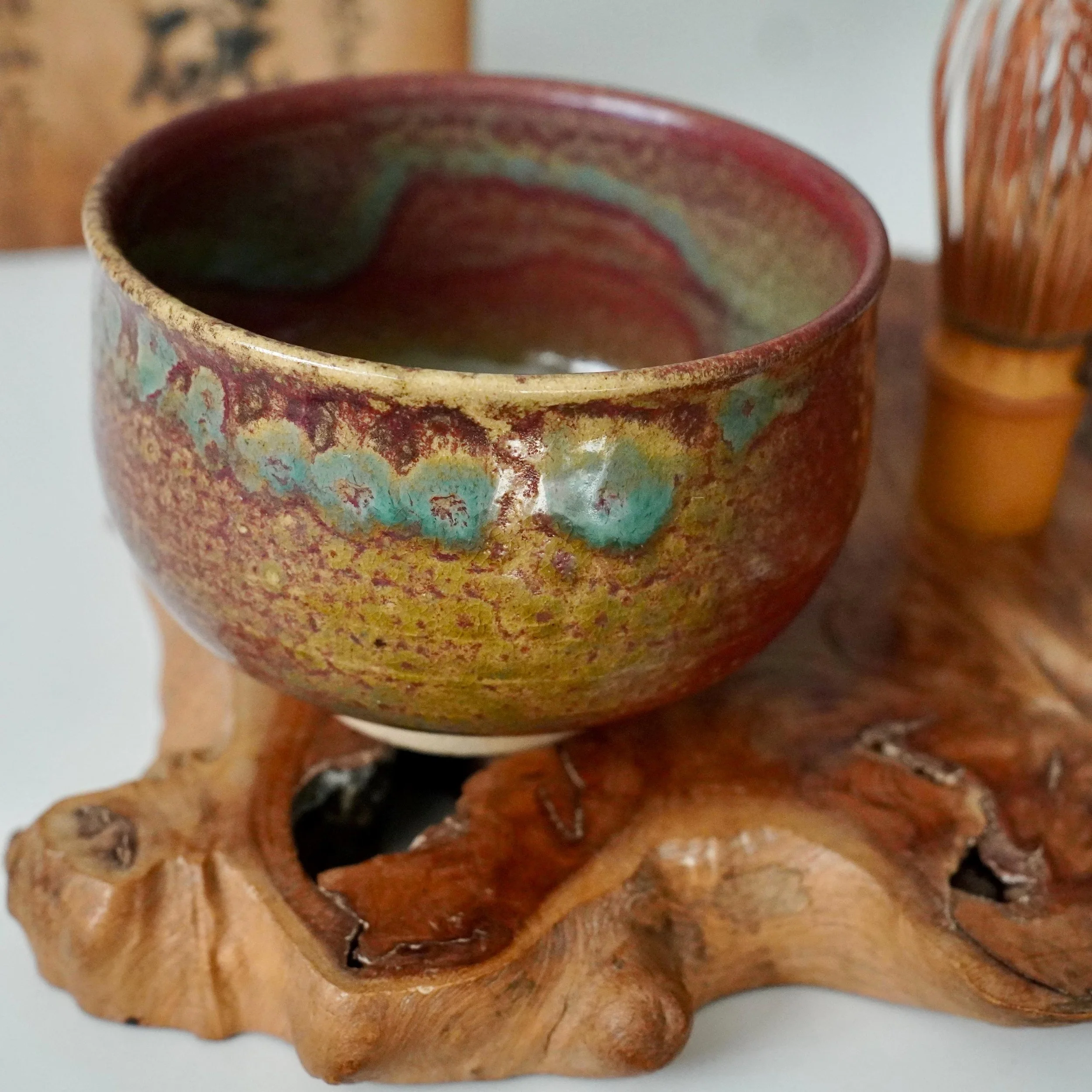 Kenzan chawan with yōhen glaze featuring ruby red, turquoise, and gold swirls, resting on a gnarled wooden stand.