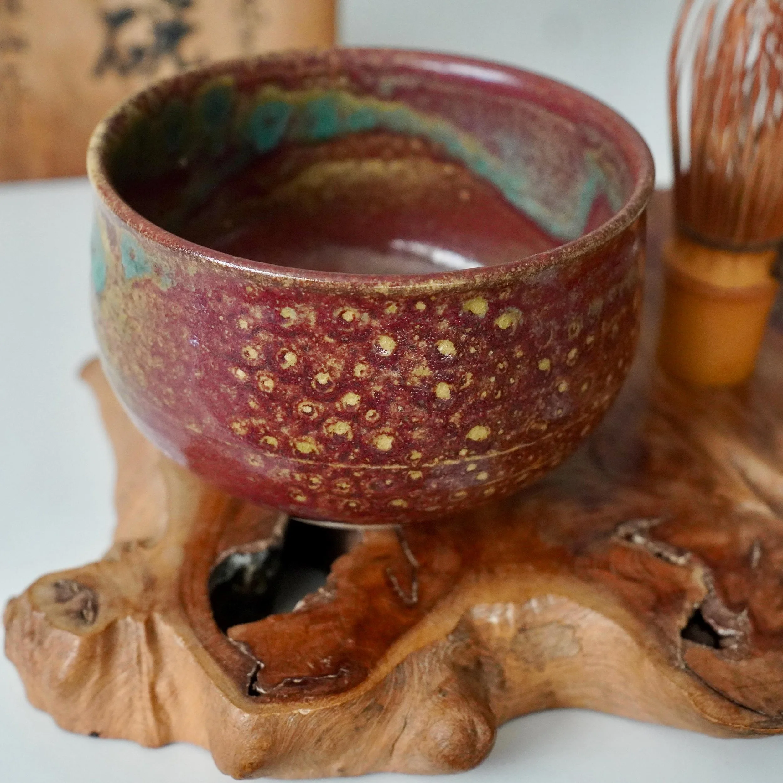 Asymmetrical ceramic chawan with a ruby red glaze pooling into turquoise and gold, resting on a carved wooden base.