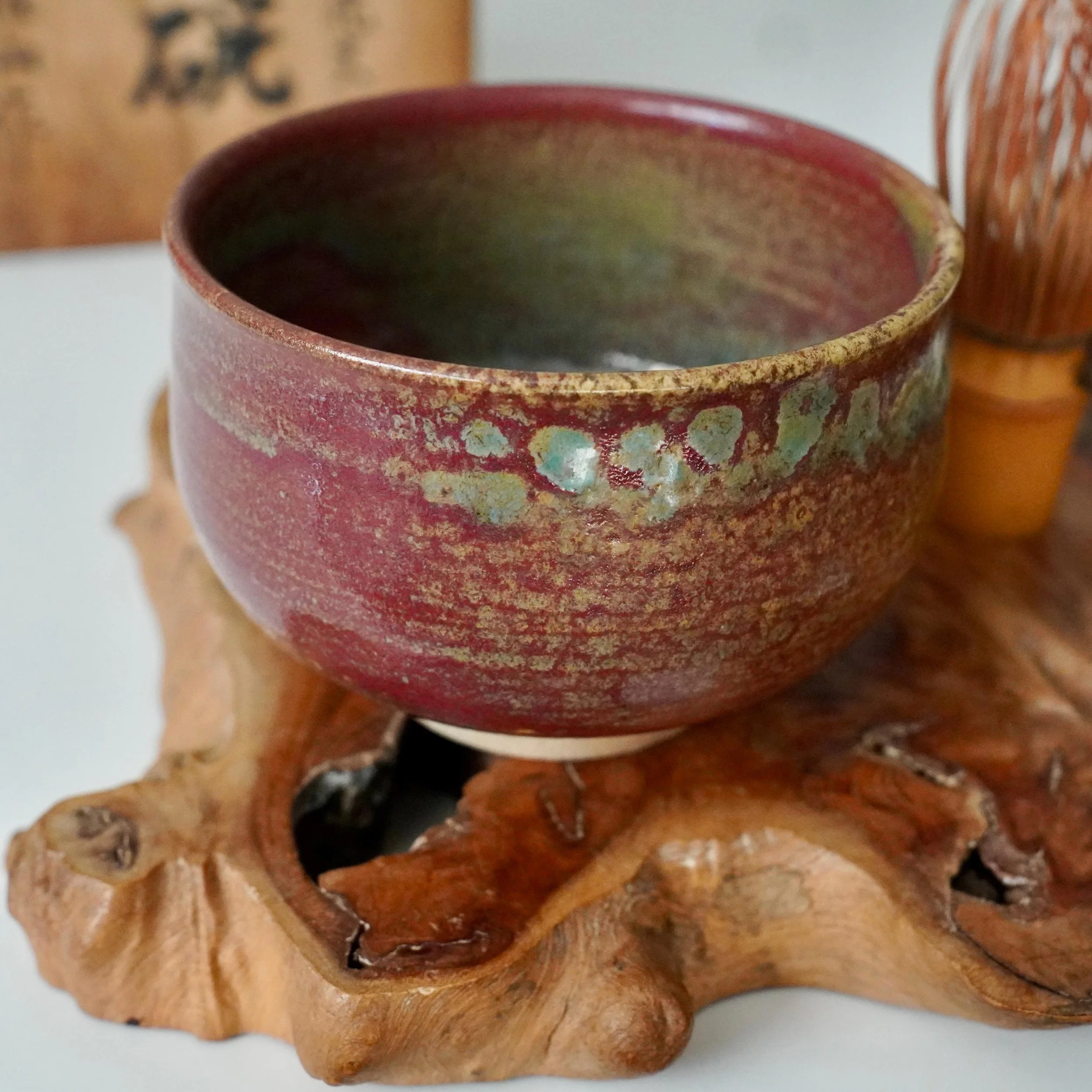 Kenzan chawan with yōhen glaze in ruby red, turquoise, and gold, on a gnarled wood stand, with a blurred chasen and tomobako in the background.