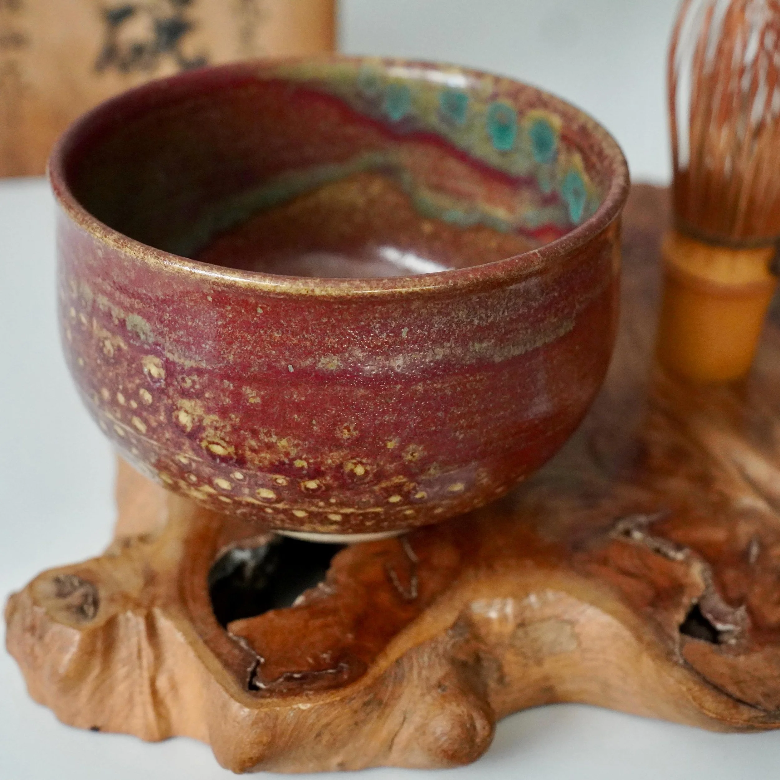 Kenzan's Yōhen glazed chawan, ruby red with yellow specks and internal turquoise dots, resting on gnarled wood. Blurred tea whisk and tomobako in background.