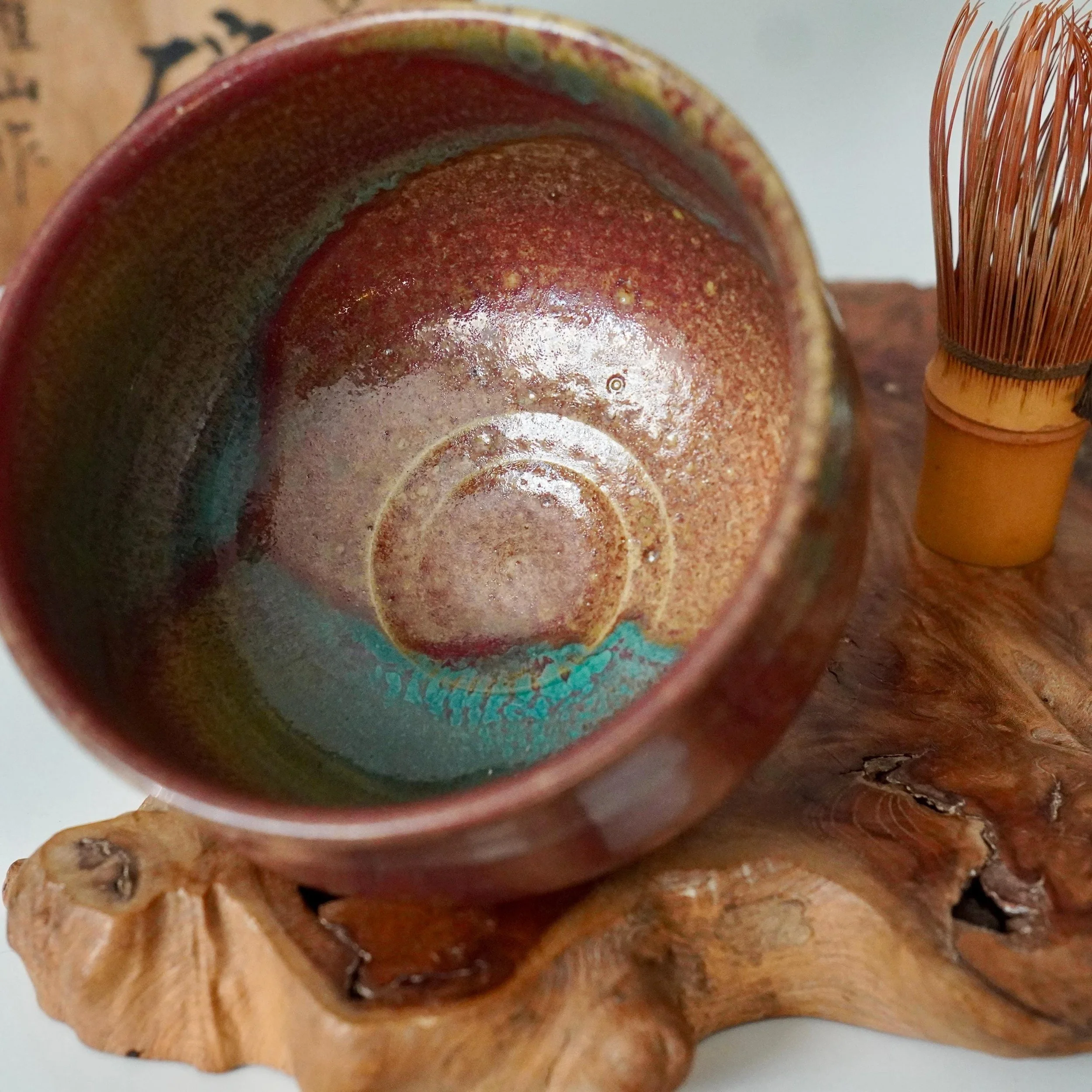 Kenzan chawan with swirling ruby red, turquoise, and gold Yōhen glaze, resting on a natural wood piece next to a bamboo whisk.