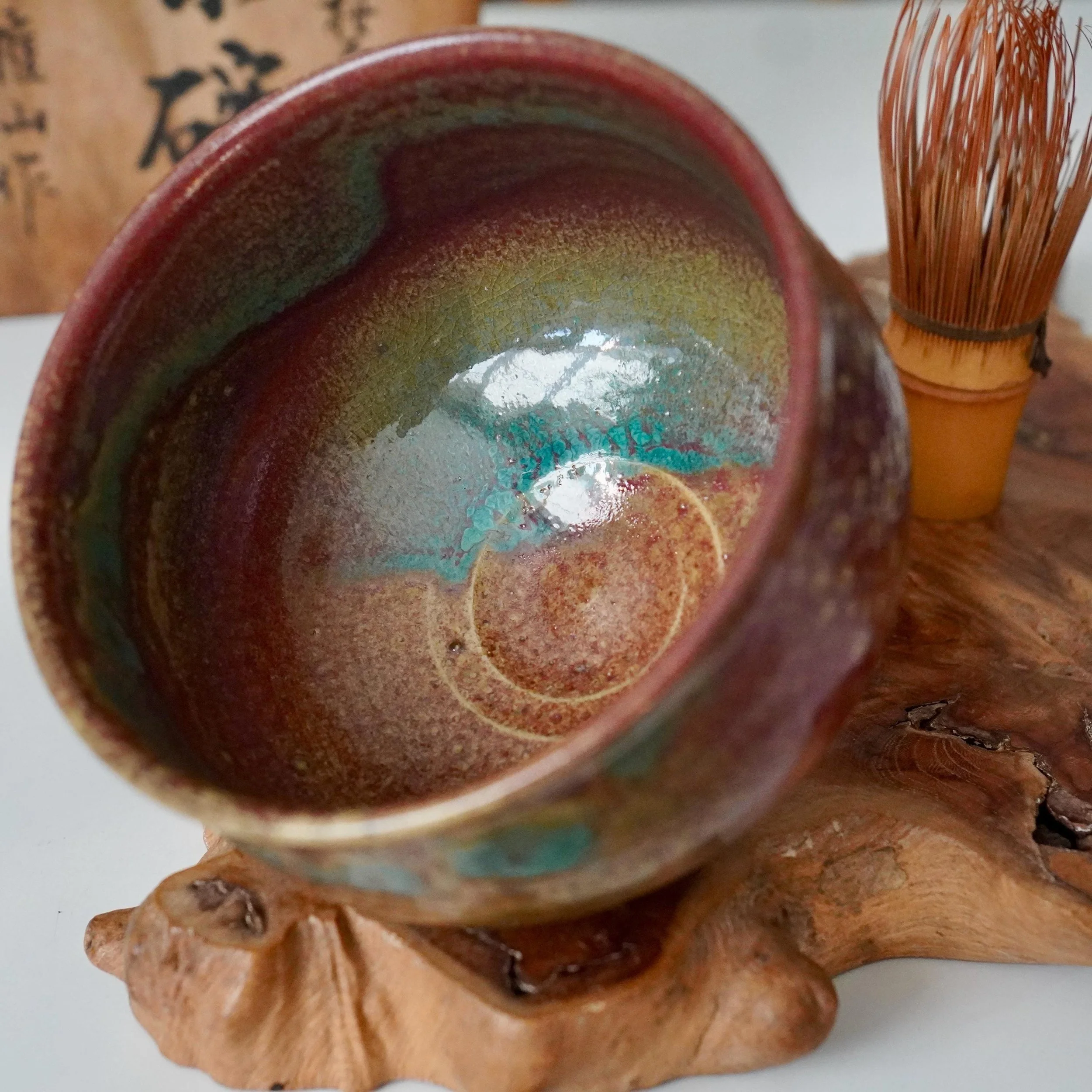 Tilted Yōhen glazed chawan by Kenzan with cracked interior of ruby red, turquoise, and gold. Bamboo whisk on a natural wood base, blurred Japanese text behind.