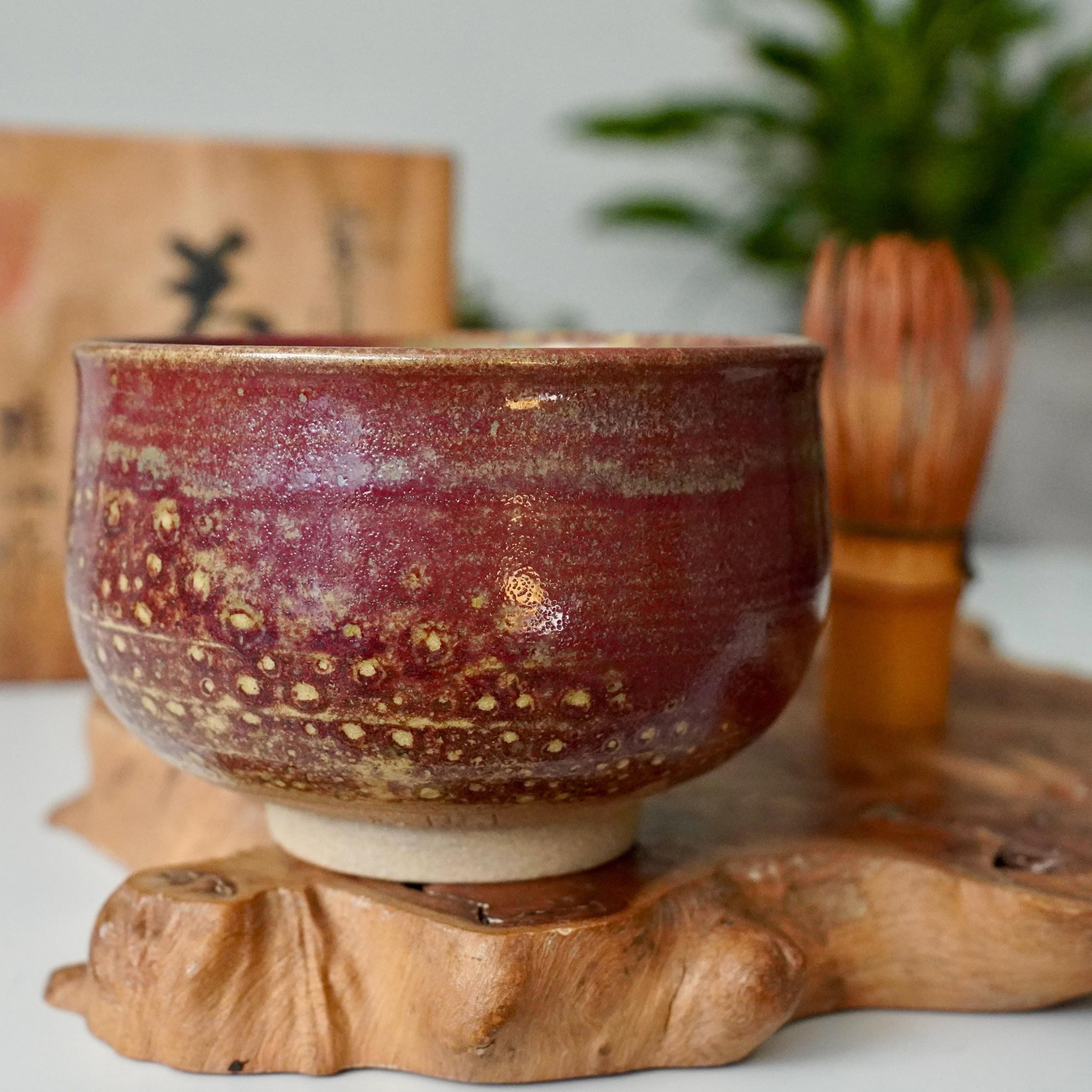 Kenzan's Yōhen glazed chawan, a deep ruby red bowl with mottled golden and subtle green-blue glaze effects, rests on a natural wood stand.