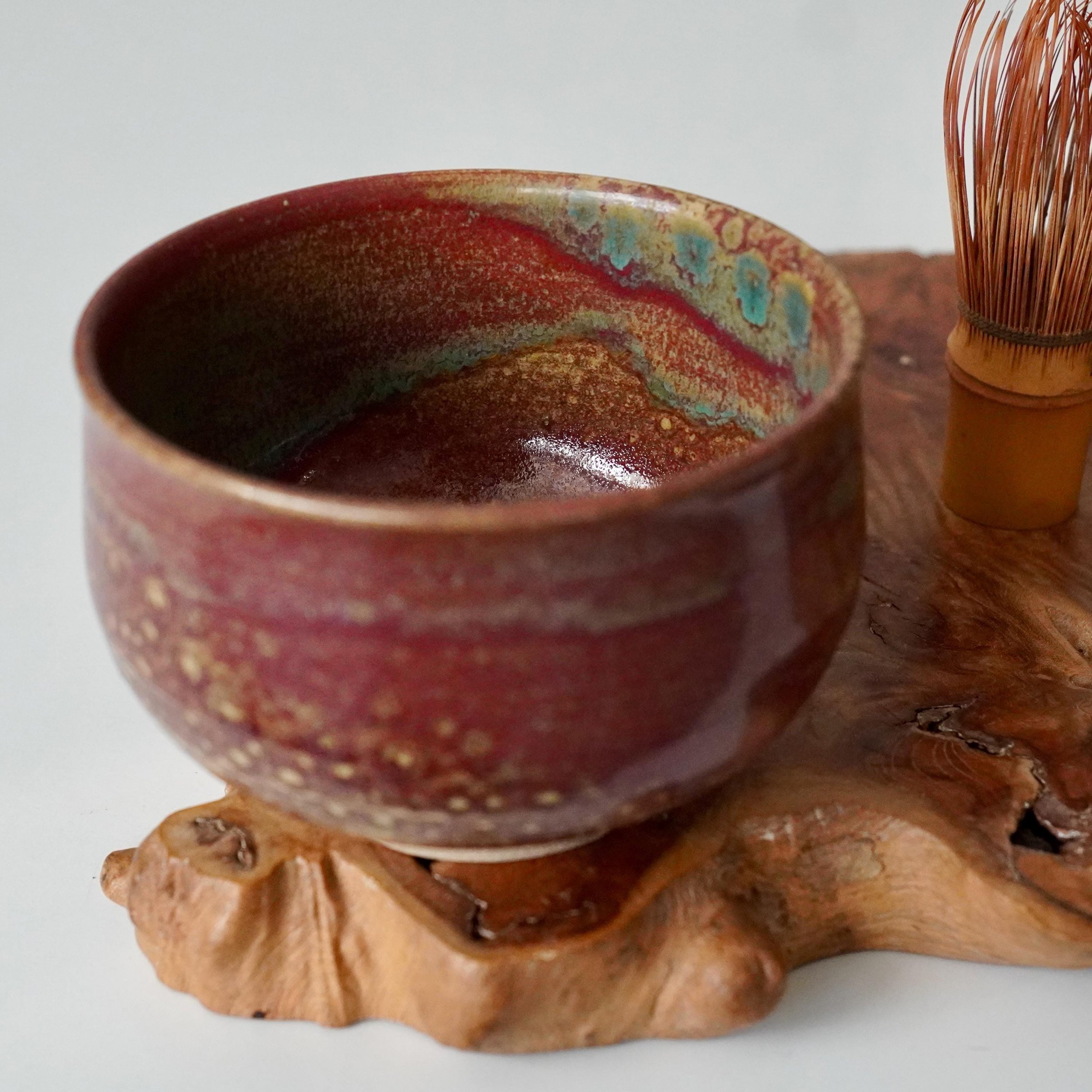 Yōhen glazed chawan by Kenzan, showing a speckled reddish exterior and a vibrant, flowing interior of ruby red, turquoise, and gold, alongside a bamboo whisk on a wooden base.