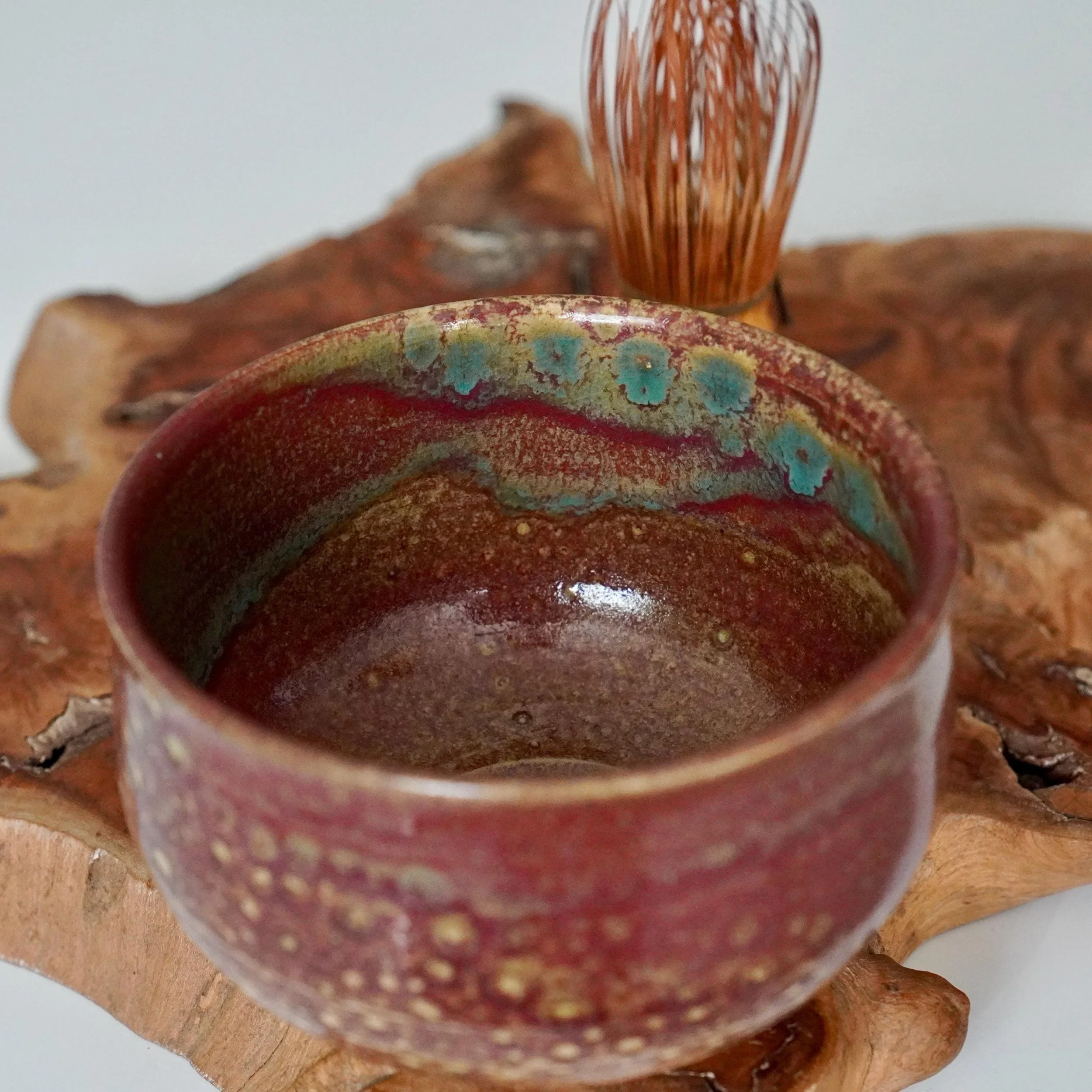 Kenzan Tokoname-yaki chawan with flowing ruby red, turquoise, and gold yōhen glaze and a subtly crackled interior, on a wood slab.