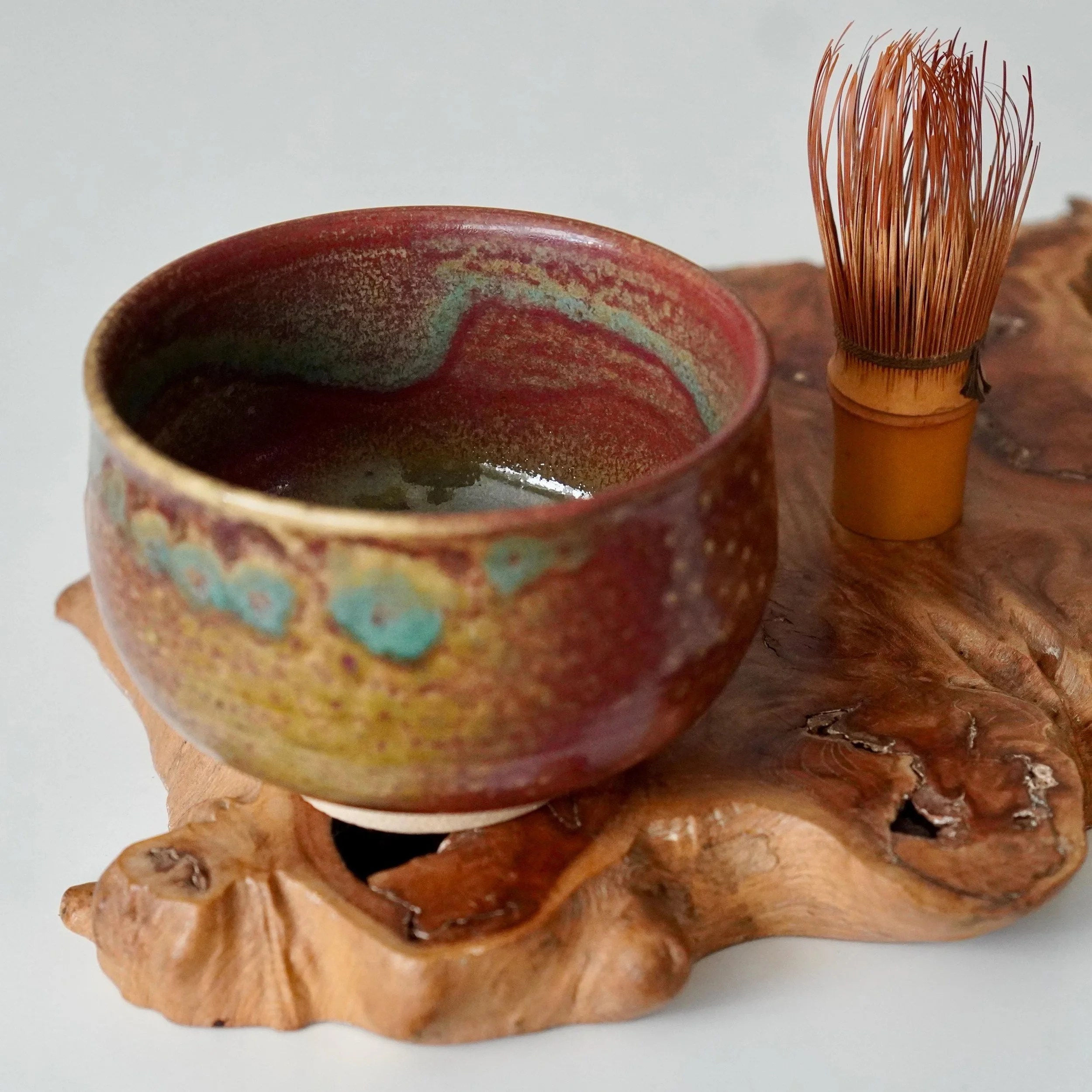 Kenzan's yōhen glazed chawan in red, turquoise, and gold, featuring an asymmetrical form. It rests on an organic wood stand next to a bamboo tea whisk.