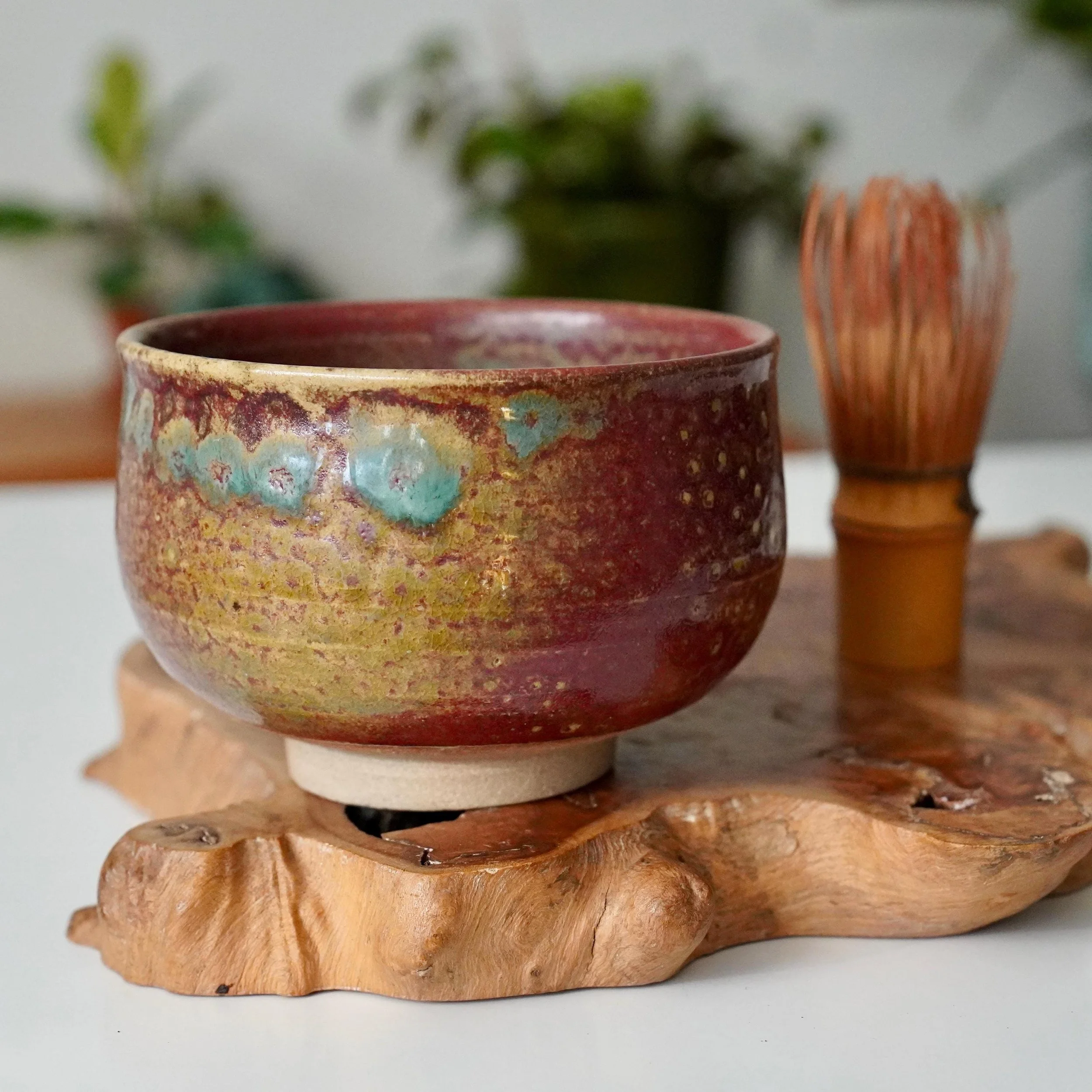 Kenzan-signed chawan with variegated Yōhen glaze in ruby red, gold, and turquoise, on a light stand atop an irregular wooden slab, with a blurred bamboo whisk.