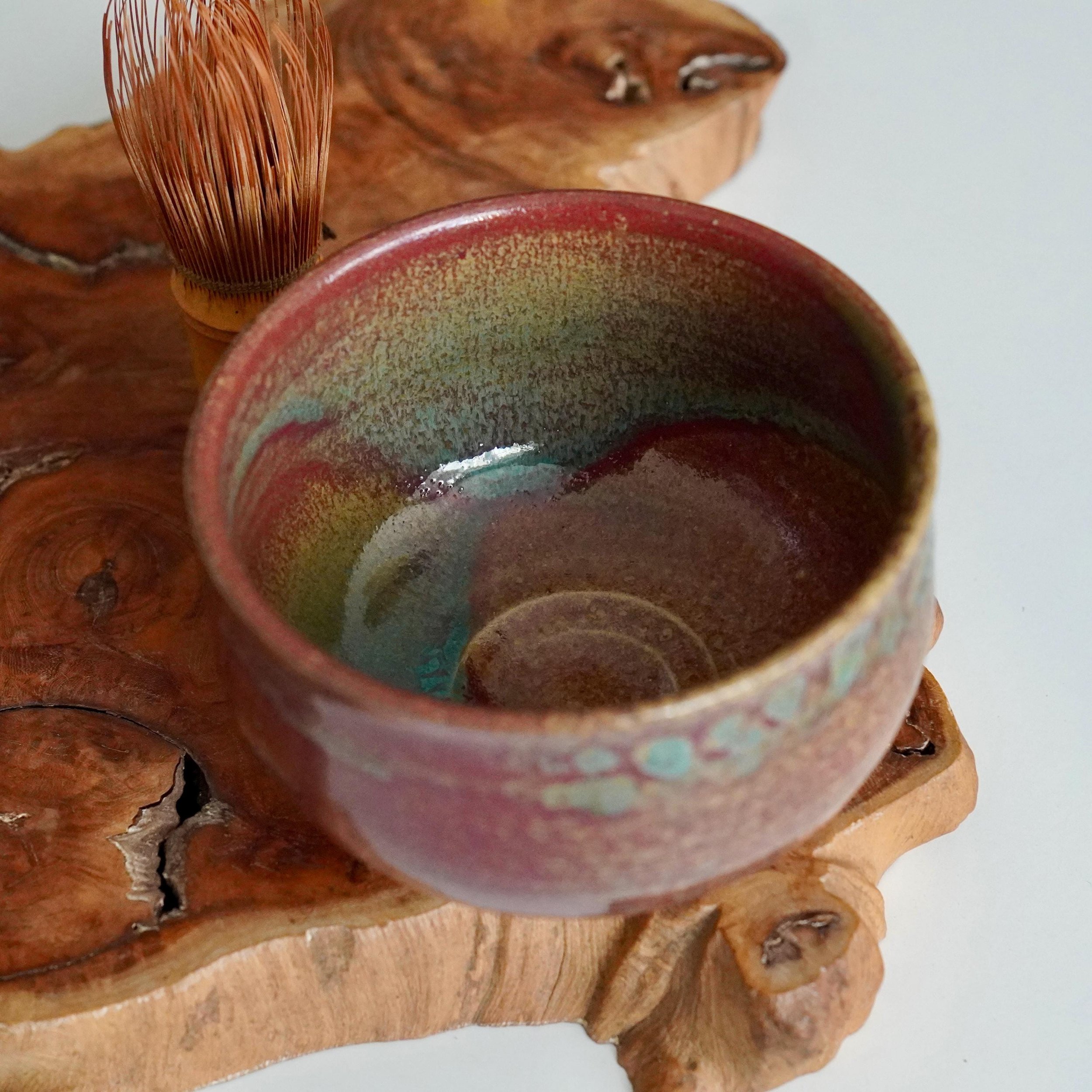 Kenzan's ceramic chawan with ruby red, turquoise, and gold yōhen glaze, showing interior crackle. A bamboo whisk rests beside it on a textured wooden slab.