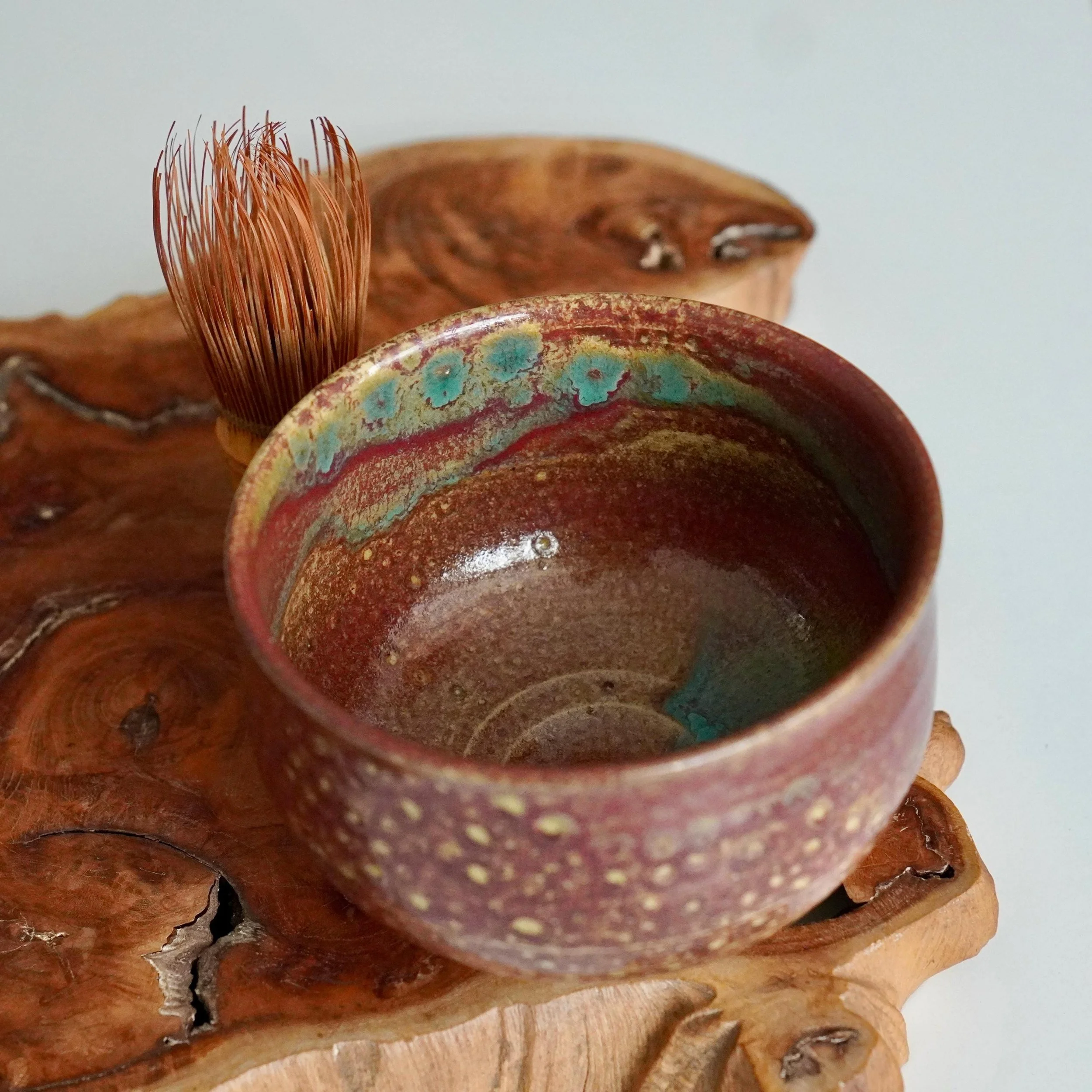 Kenzan's speckled reddish-brown chawan, interior featuring vibrant ruby, turquoise, and gold yōhen glaze. A bamboo whisk rests on rustic wood.
