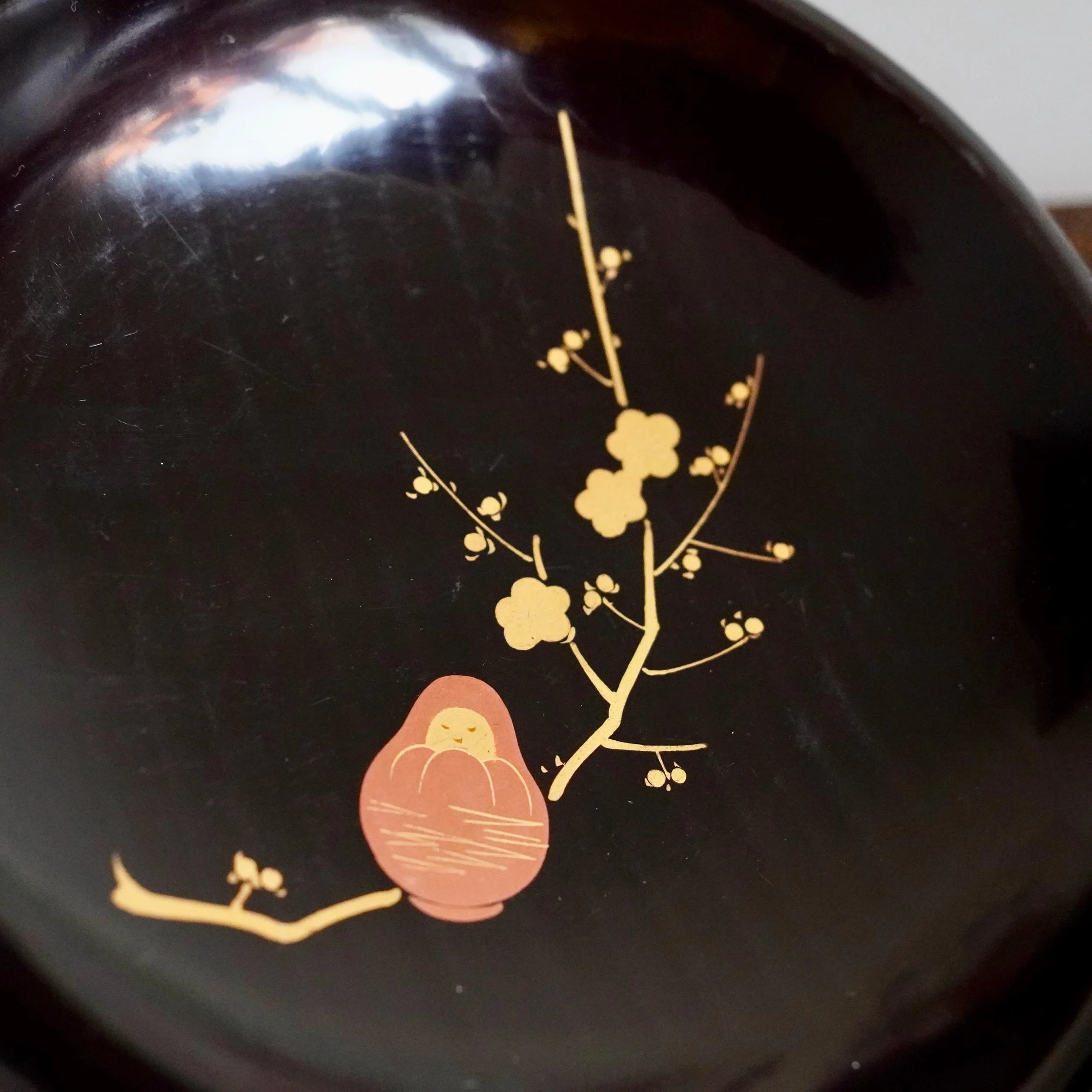 Close up of maki-e plum blossom branch and Daruma figure on black urushi wood grain visible