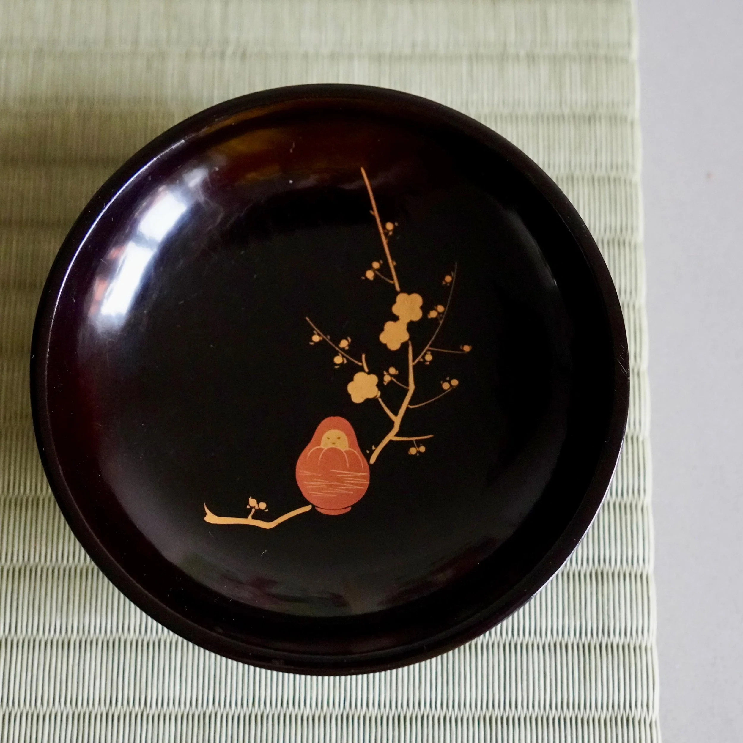 Single black urushi kashizara plate, gold plum blossom and Daruma, top view