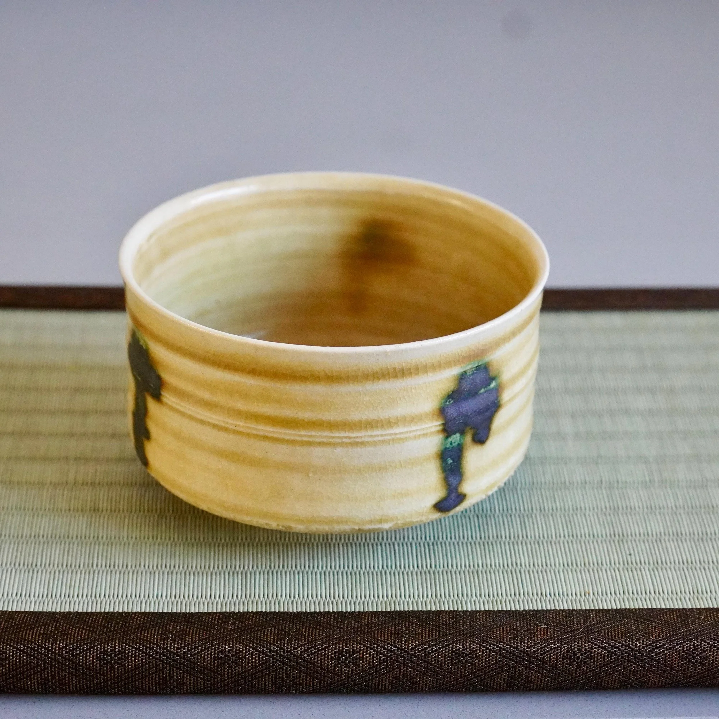 Mustard-yellow Ki-Seto chawan by Yasuda Masahiro, a ceramic tea bowl with horizontal ridges, crackled glaze, and three dark green drips, on a woven mat.