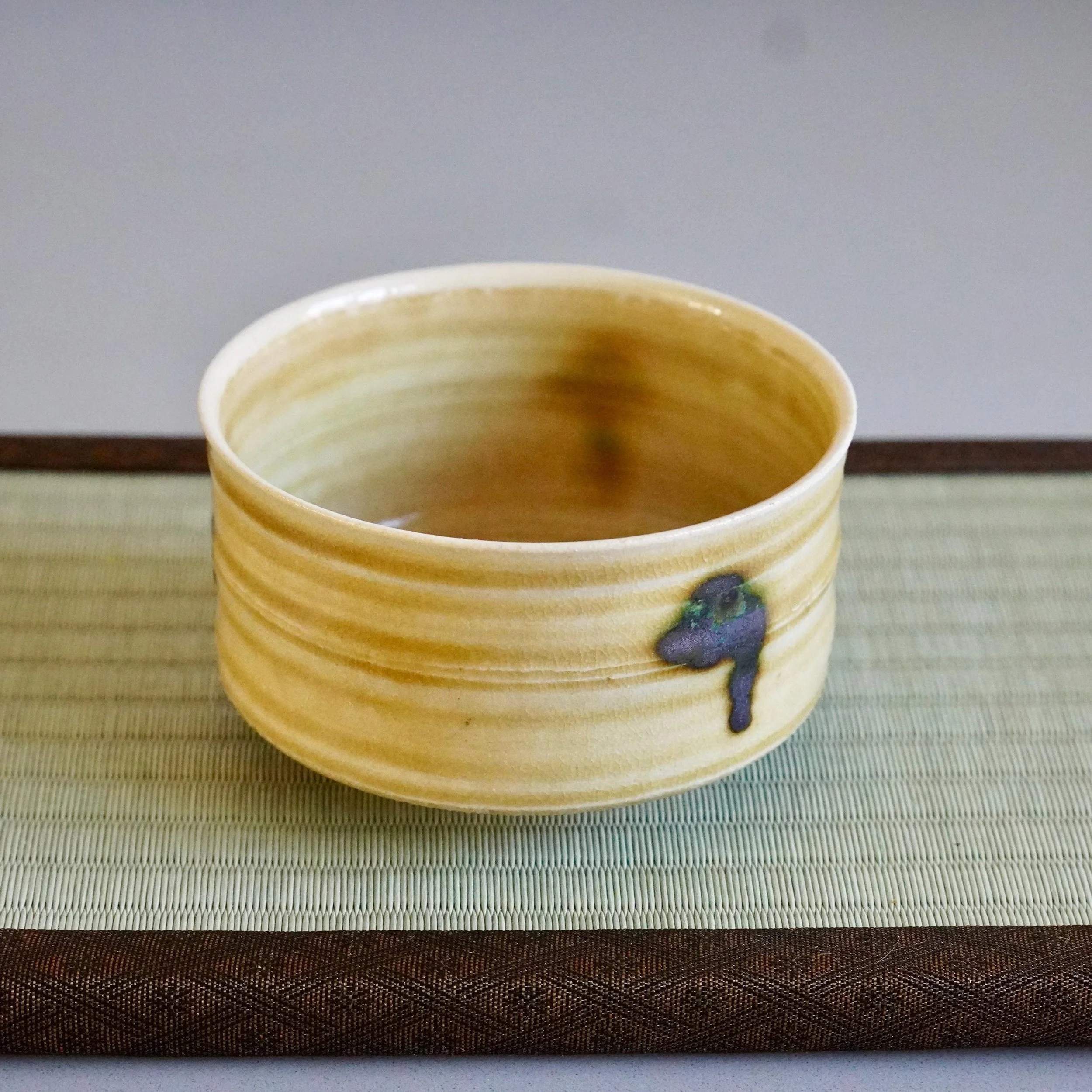 Yasuda Masahiro's mustard-yellow Ki-Seto chawan, with horizontal ridges, subtle crackle, and three green drips, resting on a tatami mat.