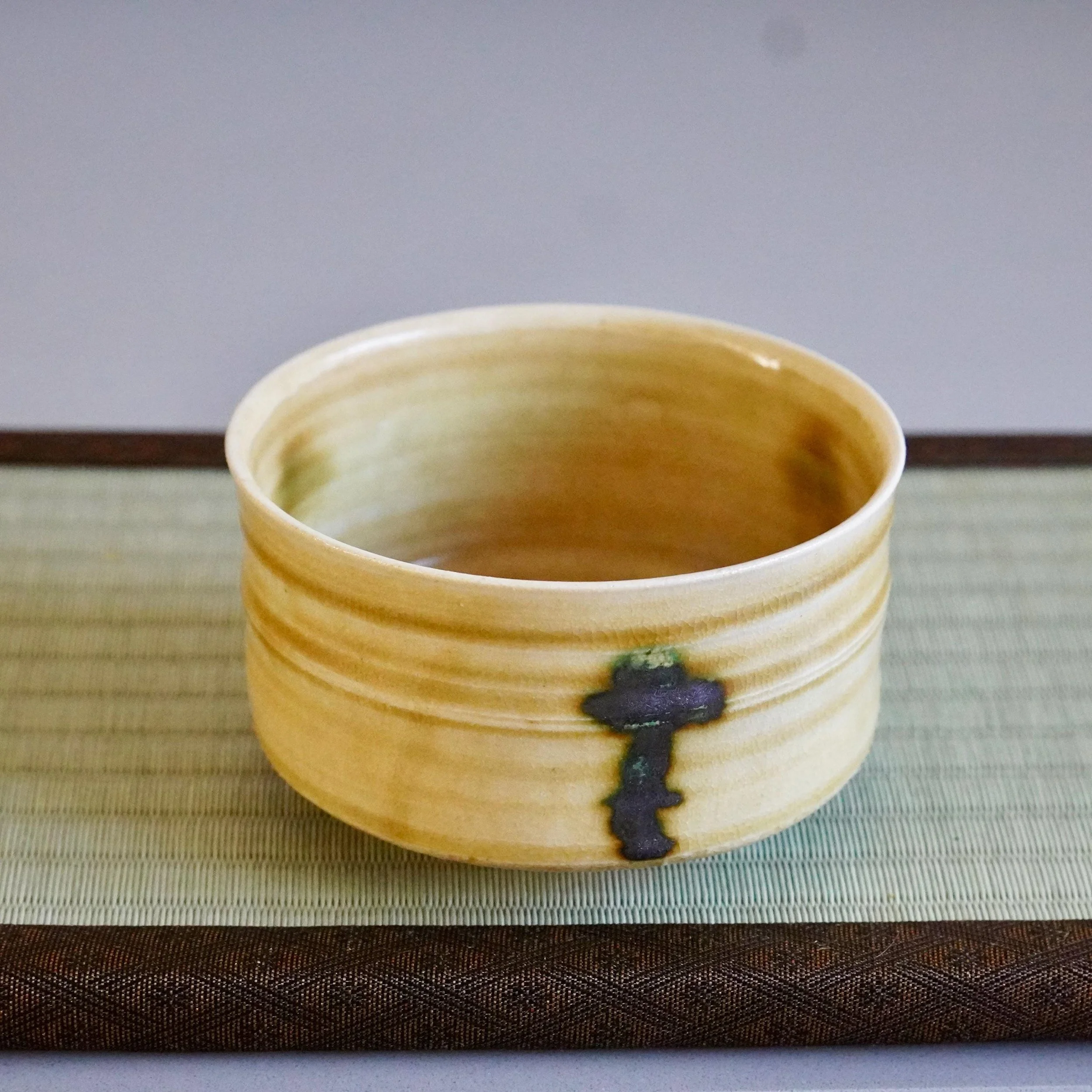 Mustard-yellow chawan by Yasuda Masahiro, with horizontal ridges and a dark blue vertical drip accented with green, sitting on a woven mat.